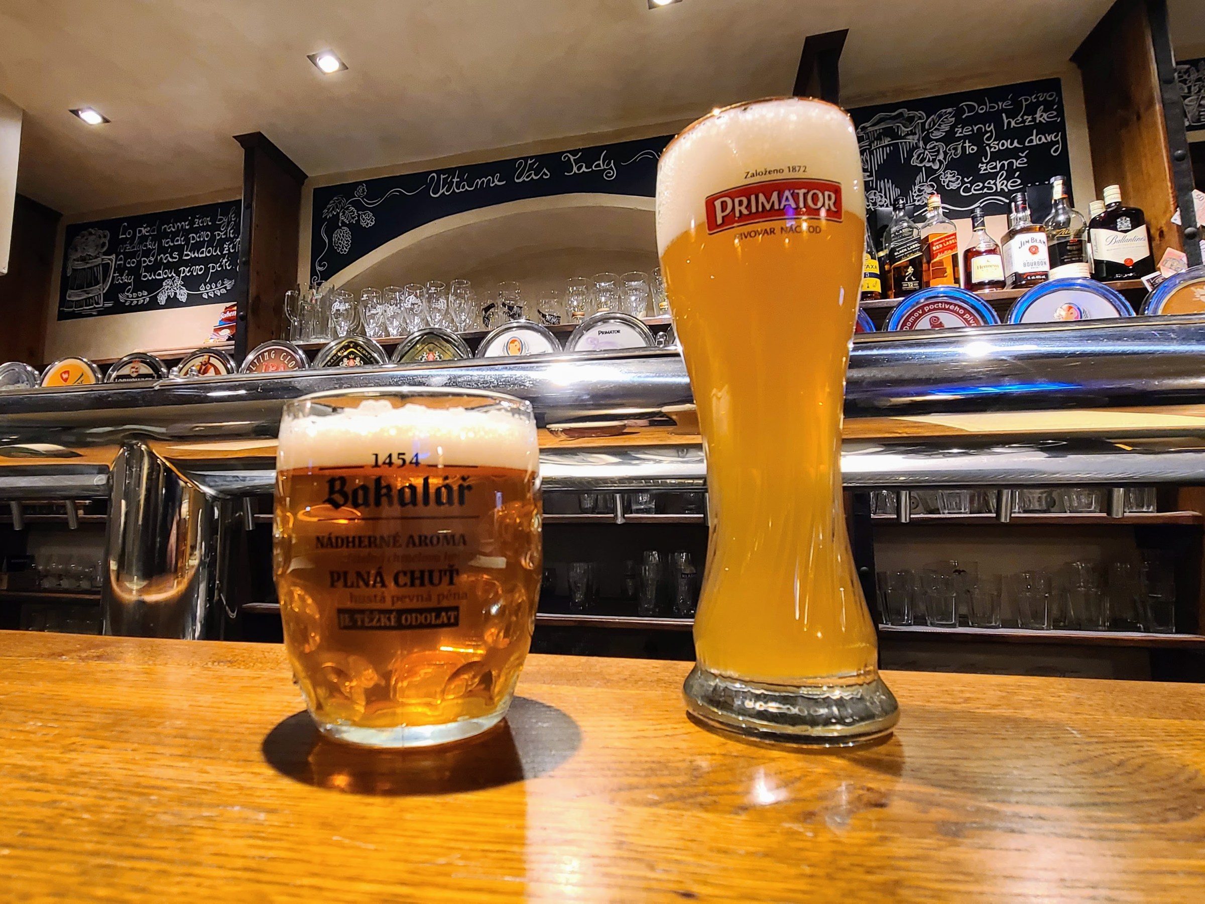 Two glasses of beer on a wooden bar with taps and shelves in the background.