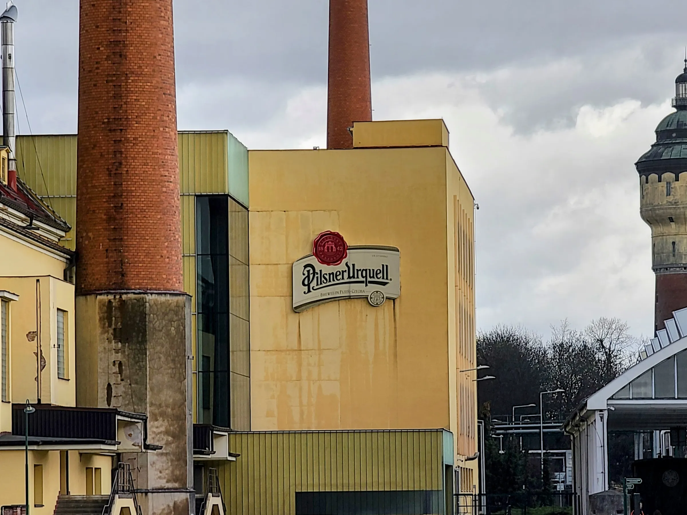 Pilsner Urquell brewery building with tall smokestacks and a tower in the background.