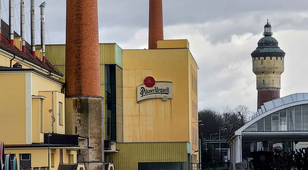 Pilsner Urquell brewery building with tall smokestacks and a tower in the background.