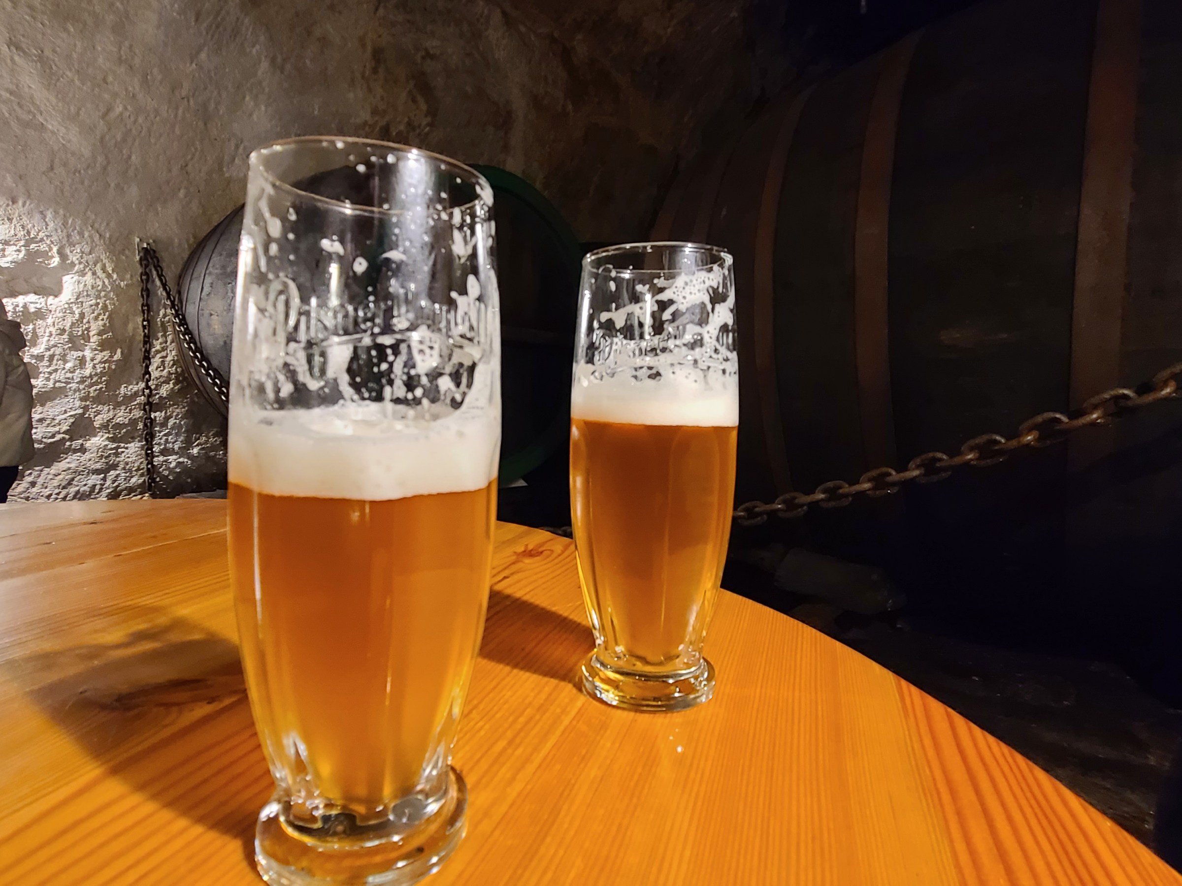 Two glasses of beer on a wooden table in a dimly lit cellar.