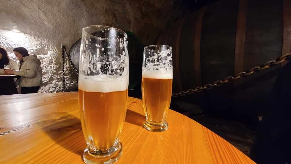 Two glasses of beer on a wooden table in a dimly lit cellar.
