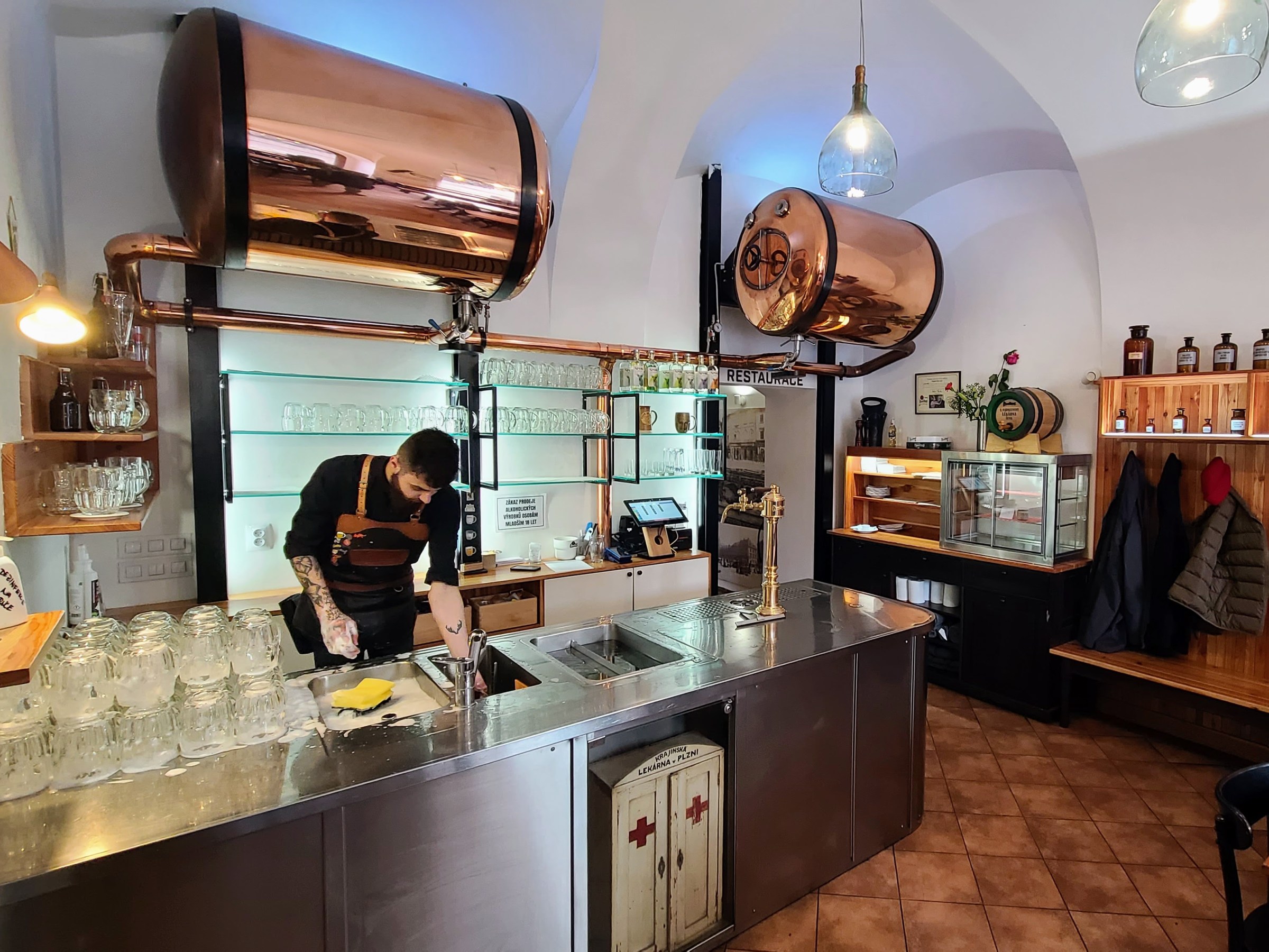 Bartender cleaning glasses at a bar with copper tanks and shelves of bottles.