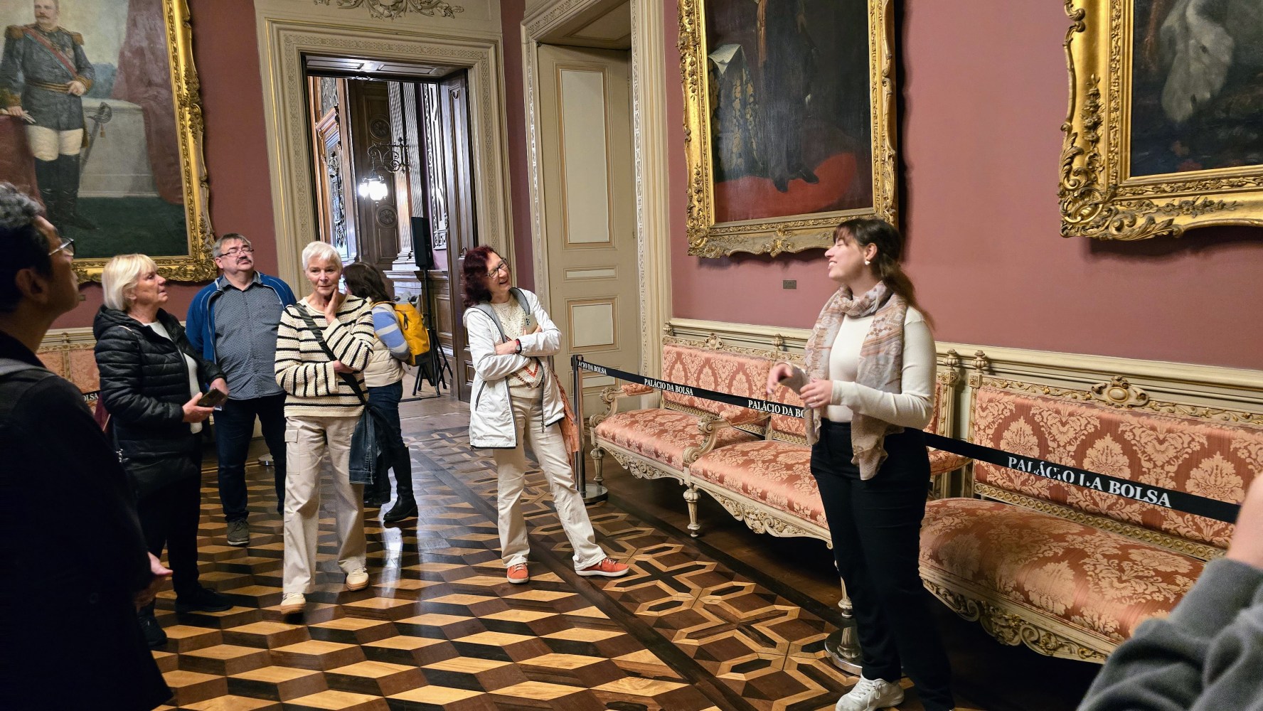 Tour guide and group in ornate gallery room with framed paintings on red walls.