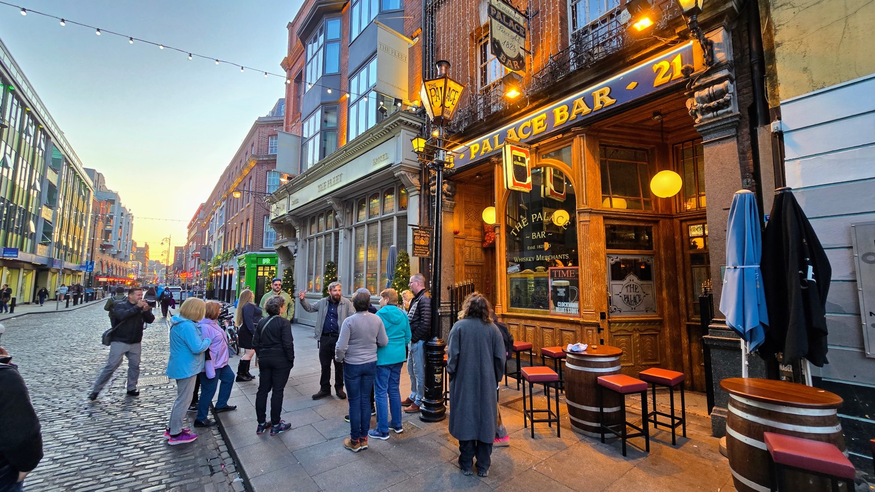 People gather outside a bar on a cobblestone street at sunset.