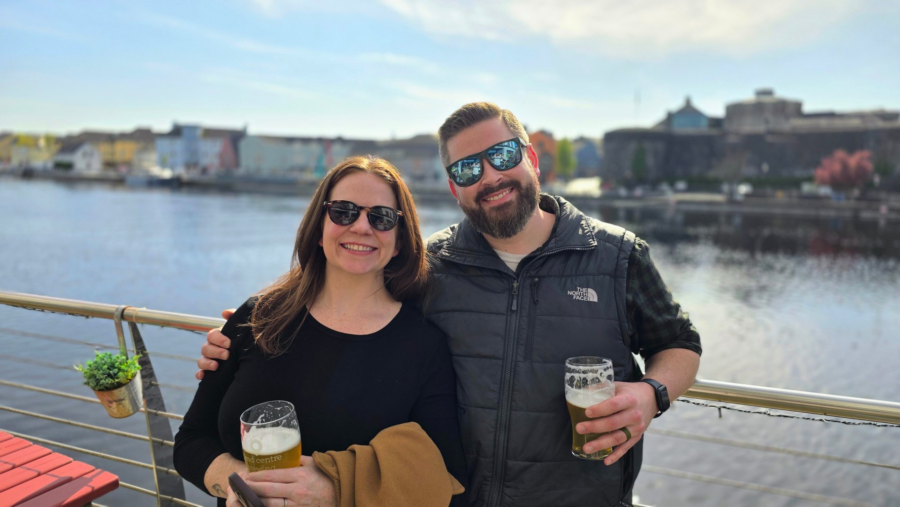 Two people in sunglasses smiling and holding beers by waterfront with buildings in background.