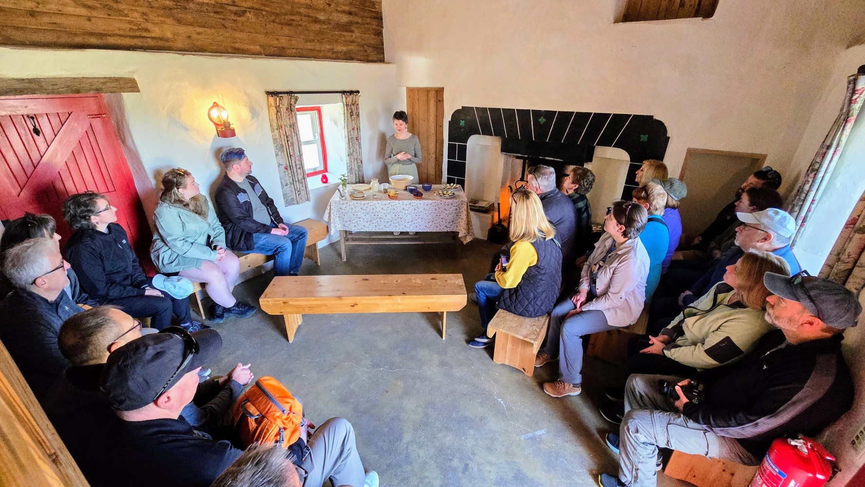 Group of people seated indoors, listening to a person standing by a table with bowls and jars.