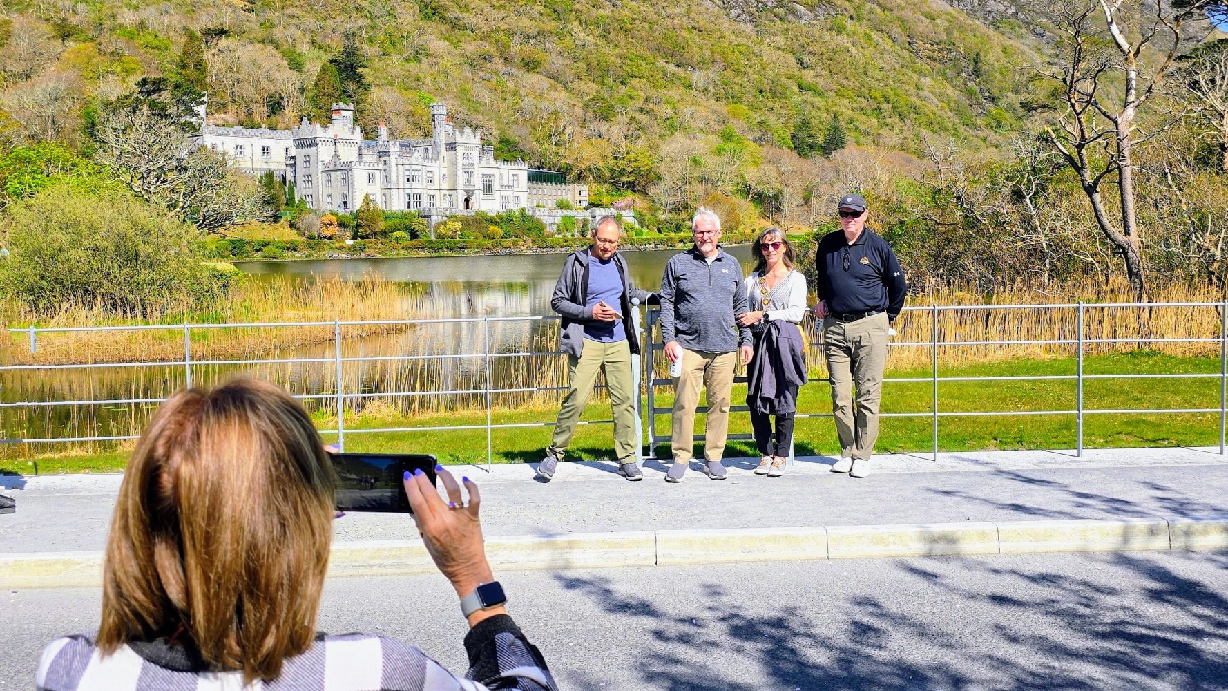 Person takes photo of four people posing in front of a scenic castle landscape.