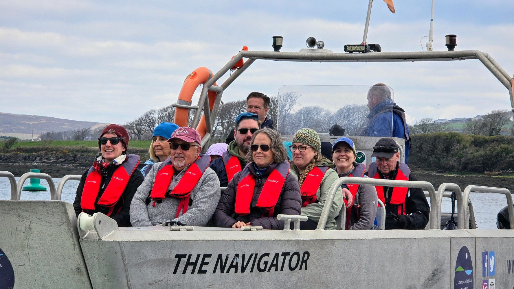 Group of people wearing red life vests on a boat named 'The Navigator' in a scenic waterway.