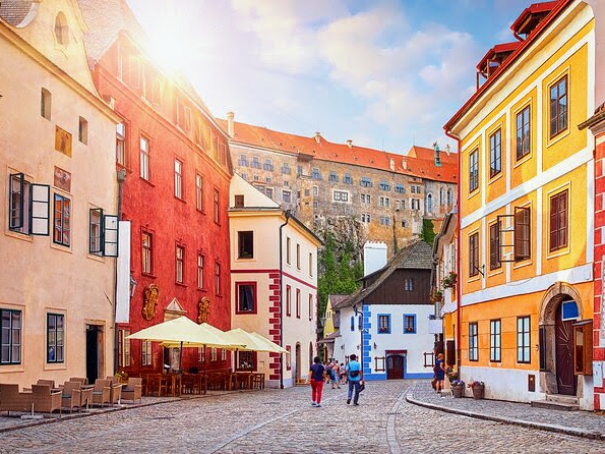 Colorful European street with historic buildings and a castle in the background under a sunny sky.