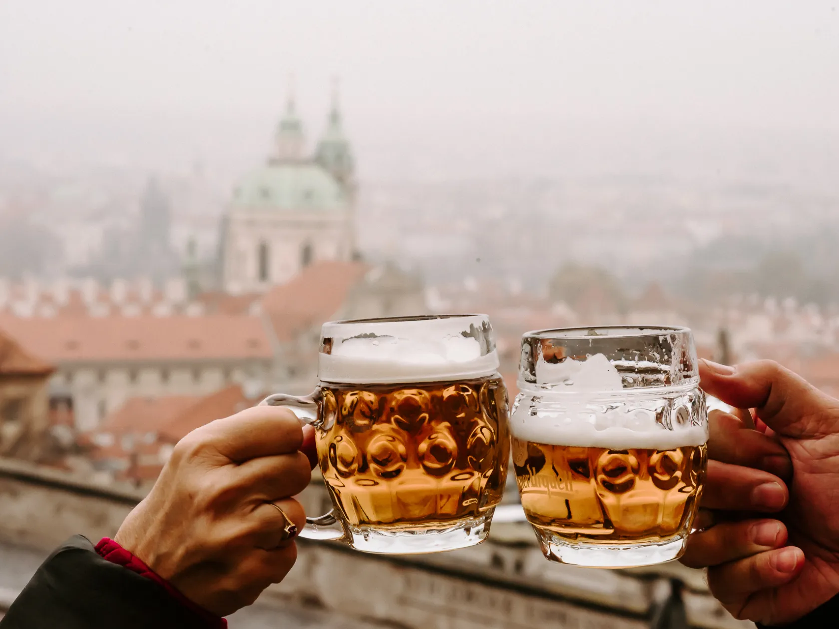Two hands holding beer mugs in cheers with a blurred cityscape in the background.