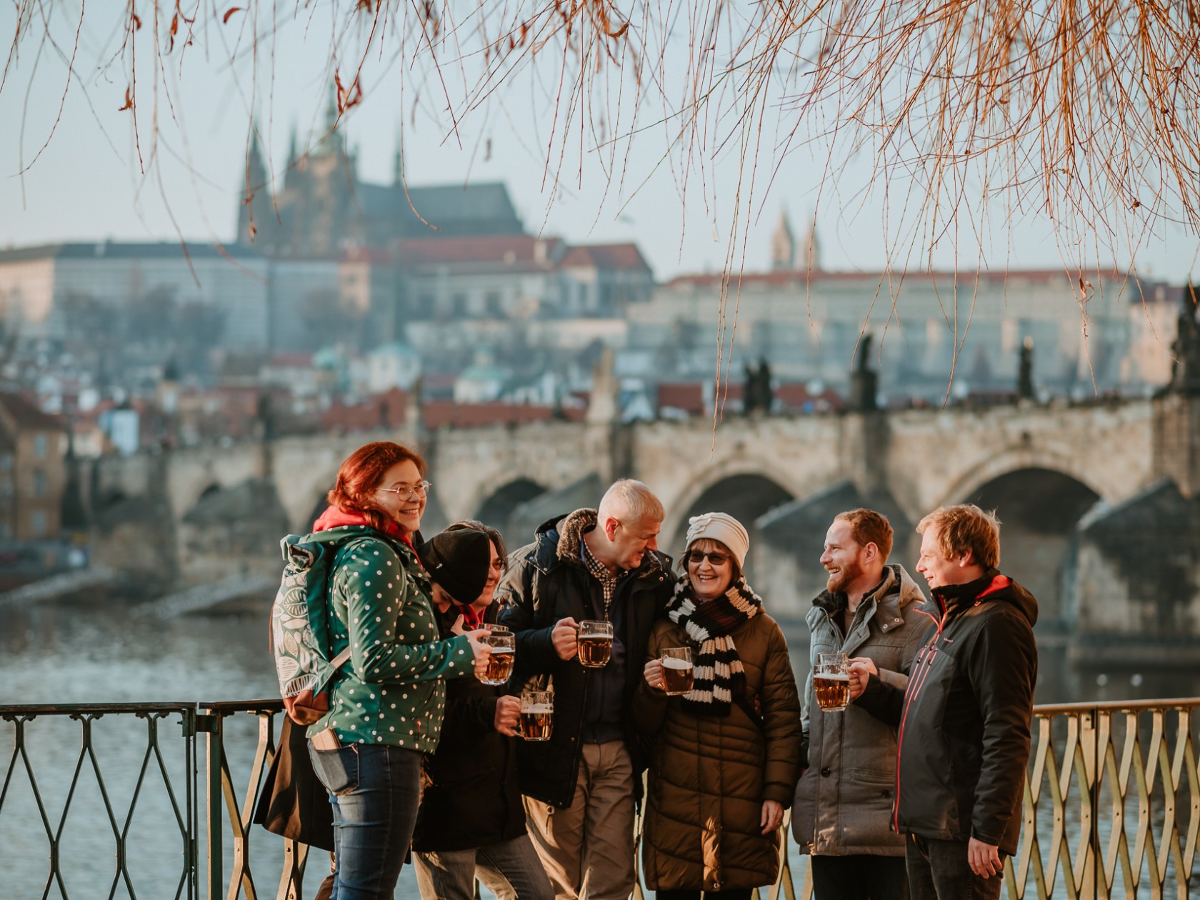 Group of people with beer mugs enjoying a riverside view near a historic bridge.