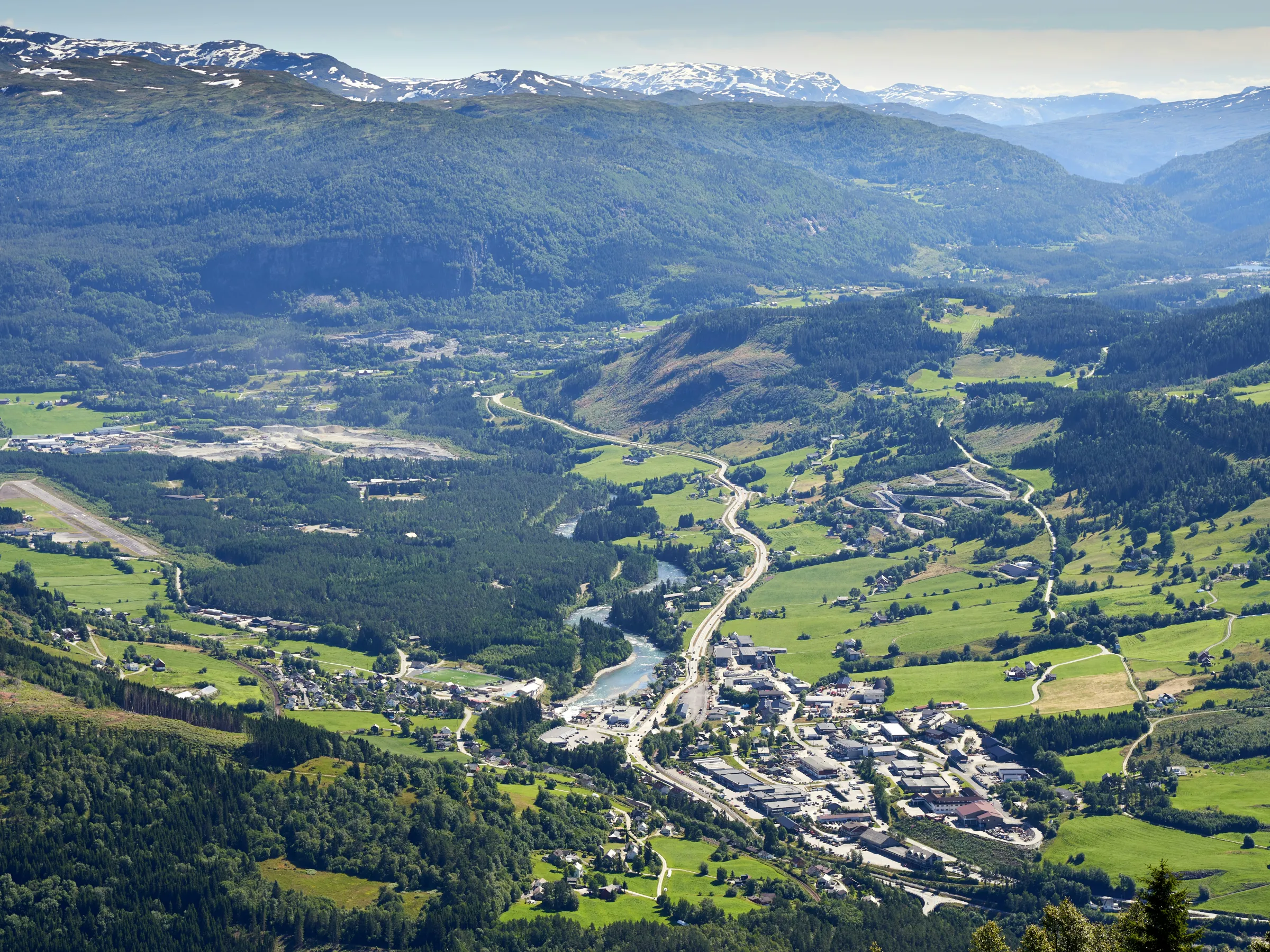 Aerial view of a valley with a river, roads, and scattered buildings surrounded by green hills and distant mountains.
