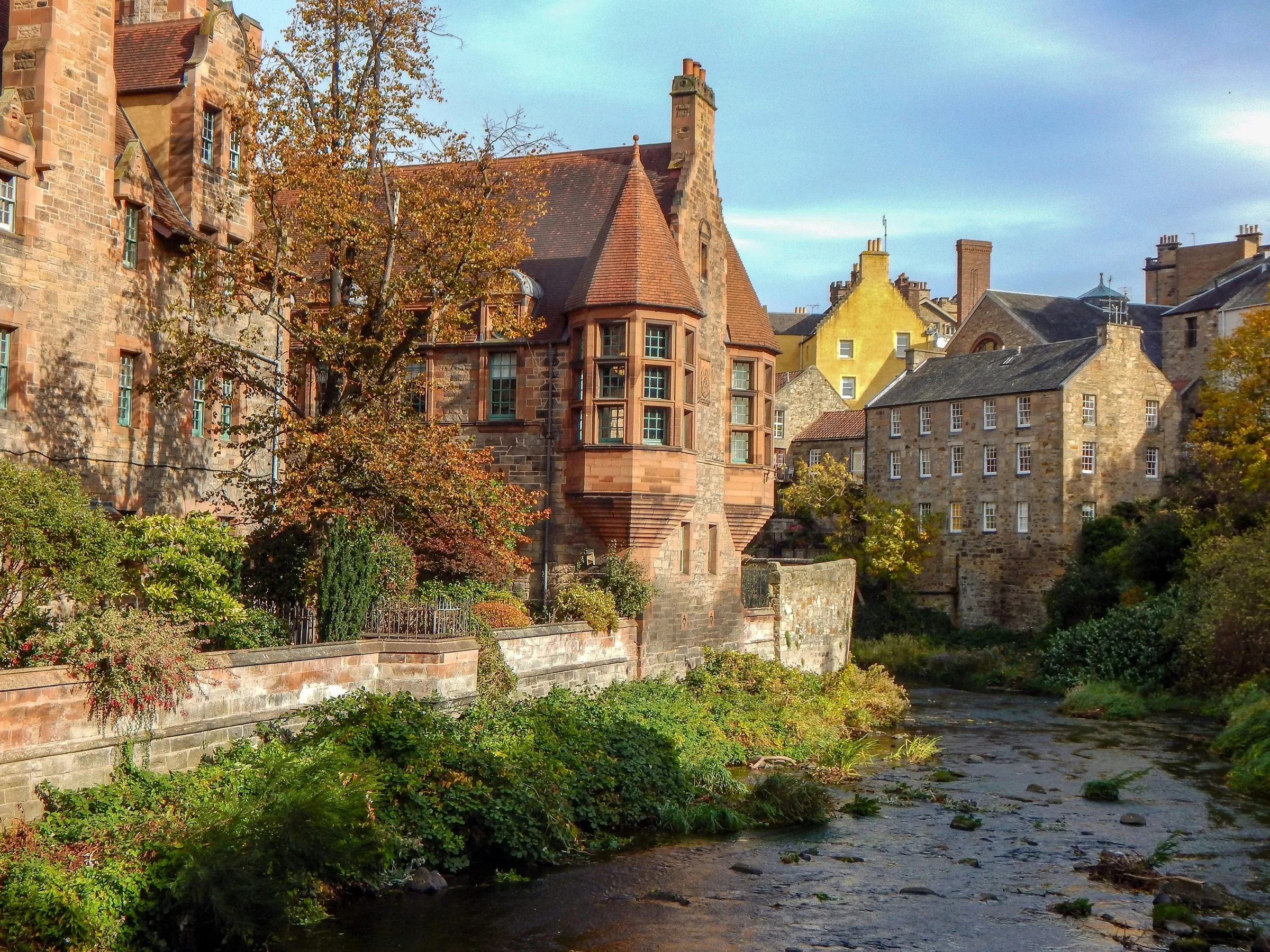 Historic stone buildings by a tree-lined river under a clear sky.