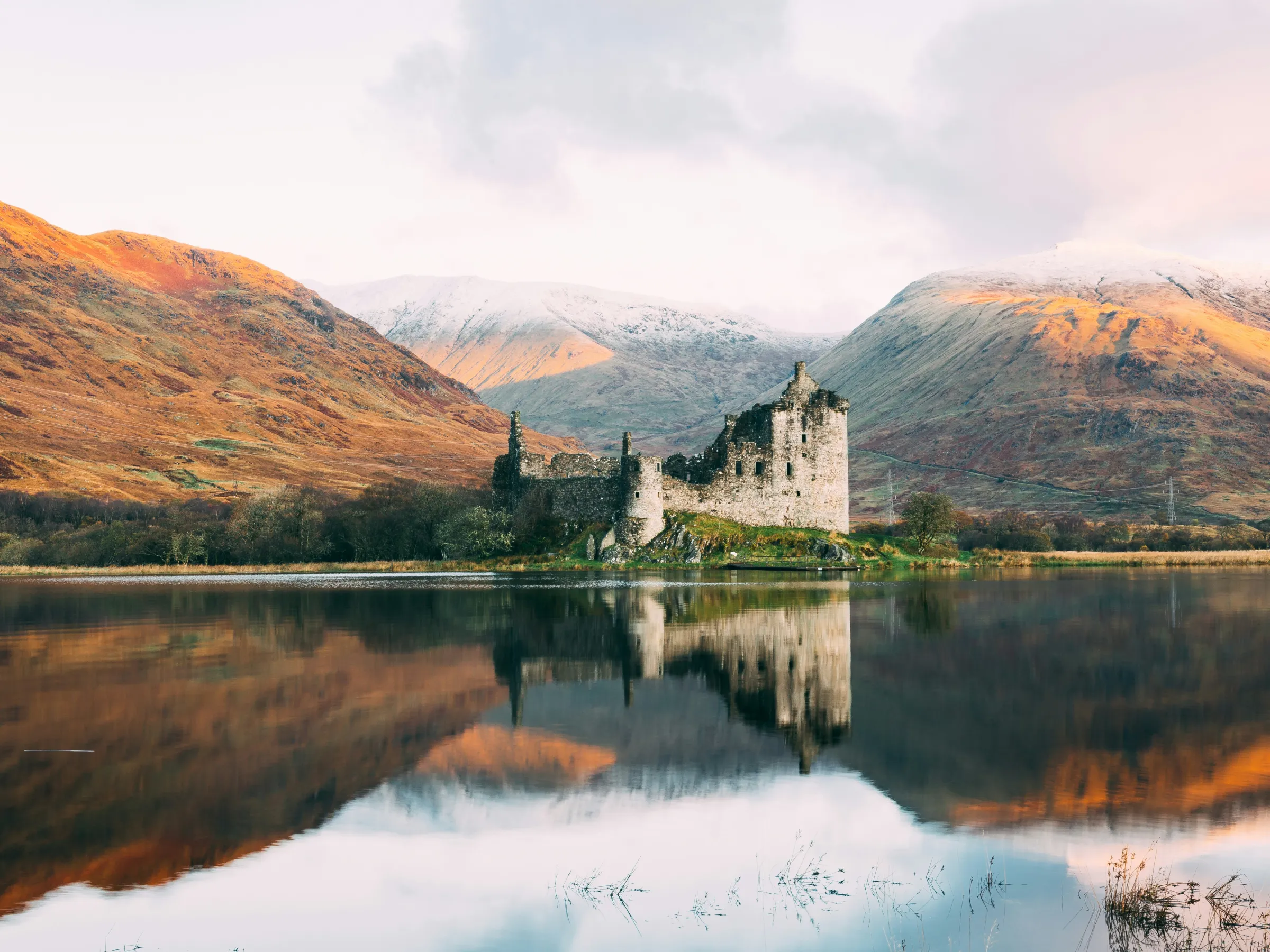 Ruined castle reflected in a lake with mountainous background at sunrise.