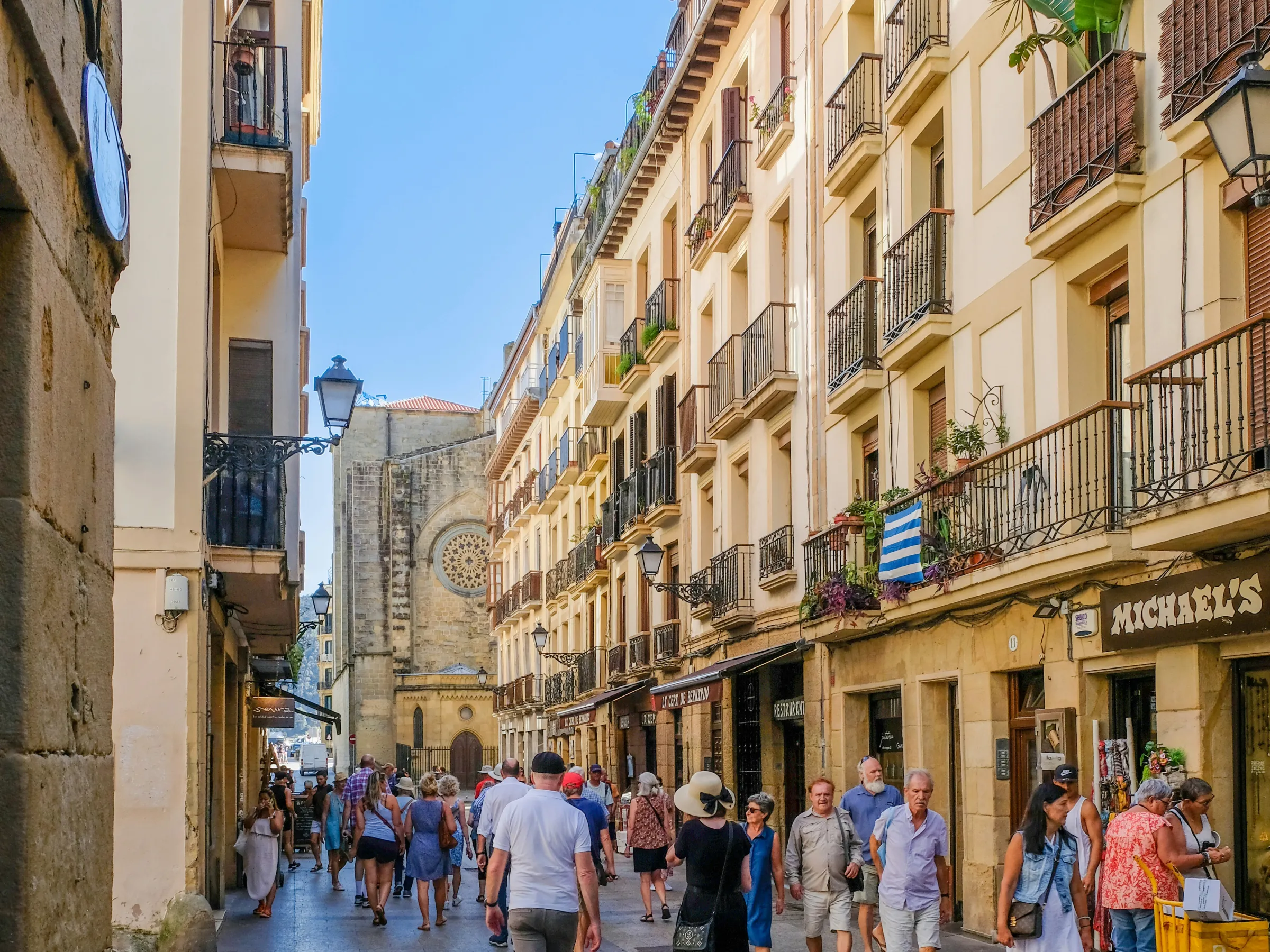 People walk along a narrow street with old buildings and balconies on a sunny day.