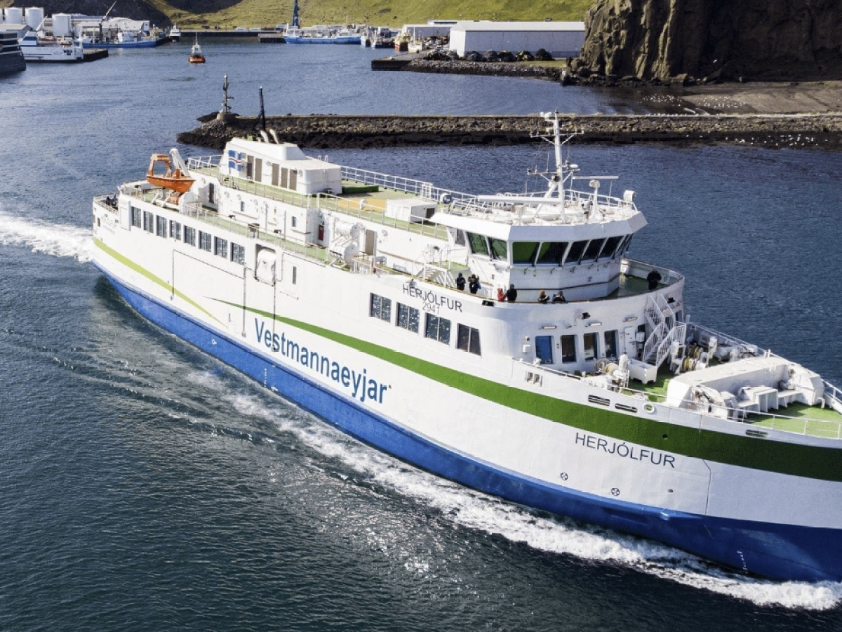 Large ferry named Herjólfur sailing in a harbor with industrial buildings and hills in the background.