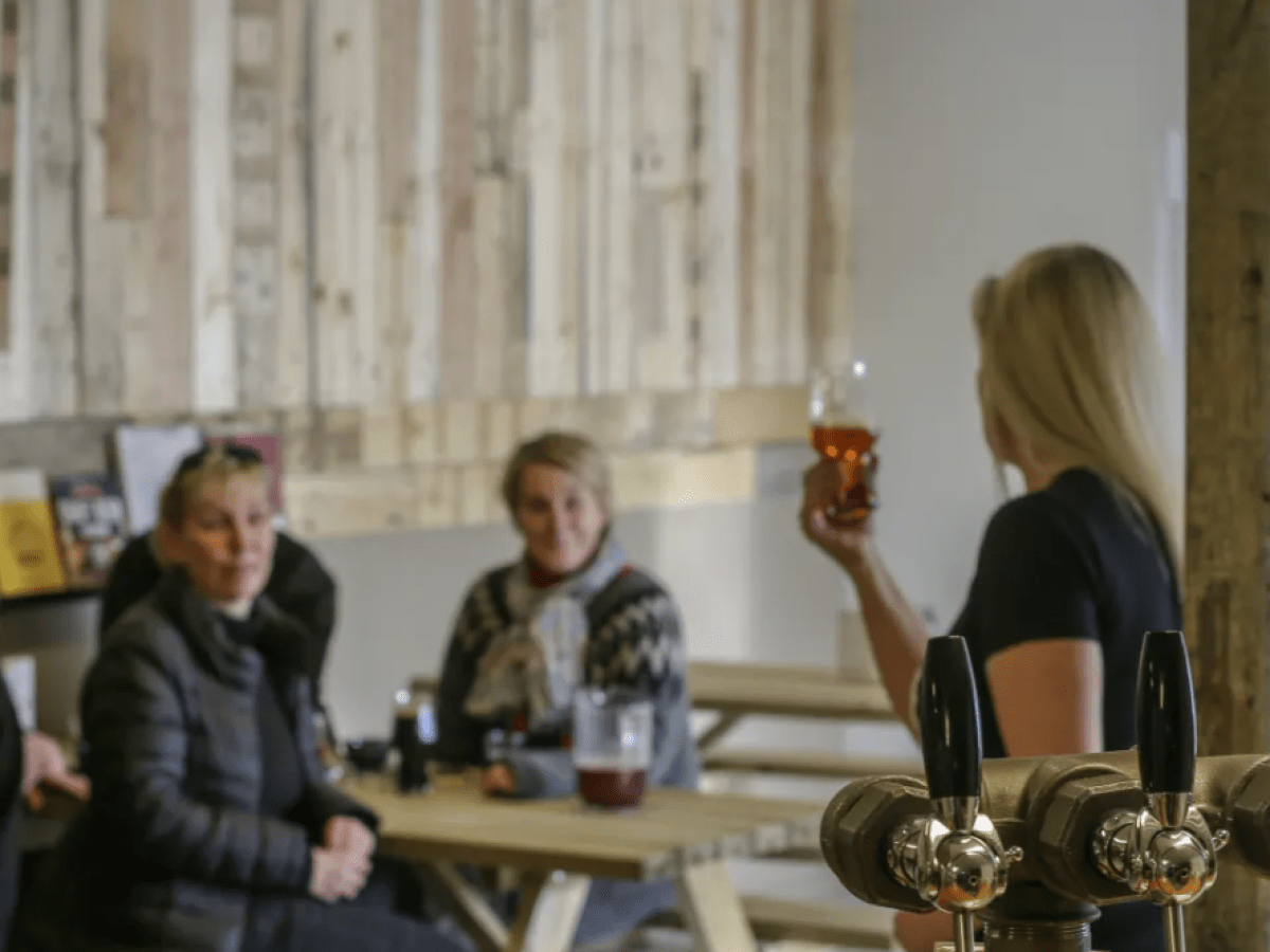 Woman raising glass in front of a seated group in a rustic room with beer taps.