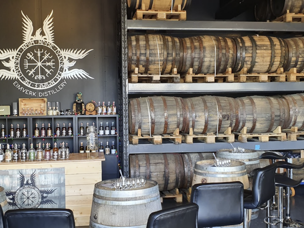 Distillery interior with barrels on shelves, bottles on countertop, and tasting tables with stools.