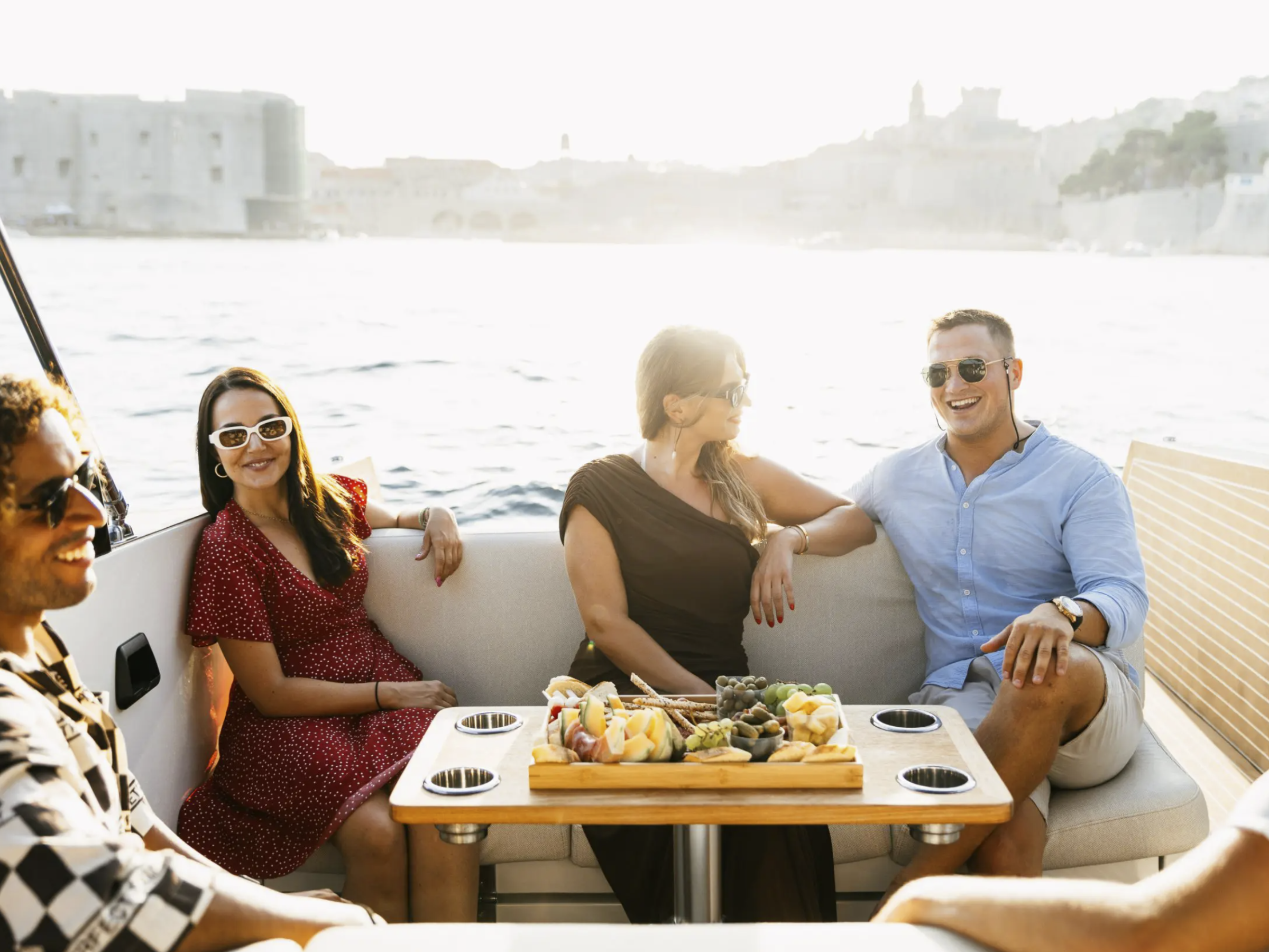 Group of friends laughing on a boat, enjoying snacks with a scenic view in the background.