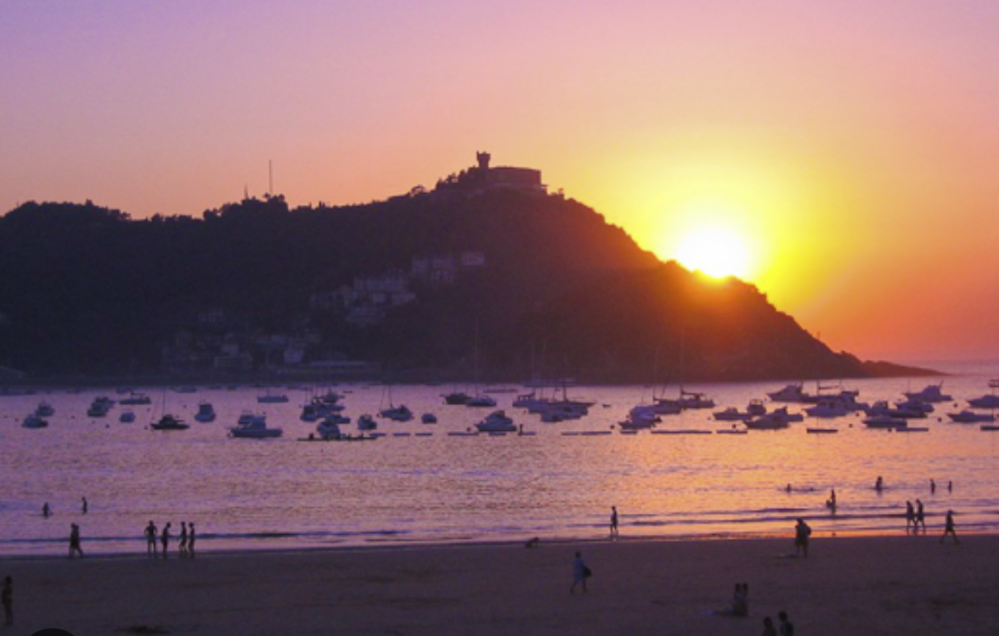 Sunset over a bay with boats and silhouettes of people on the beach.