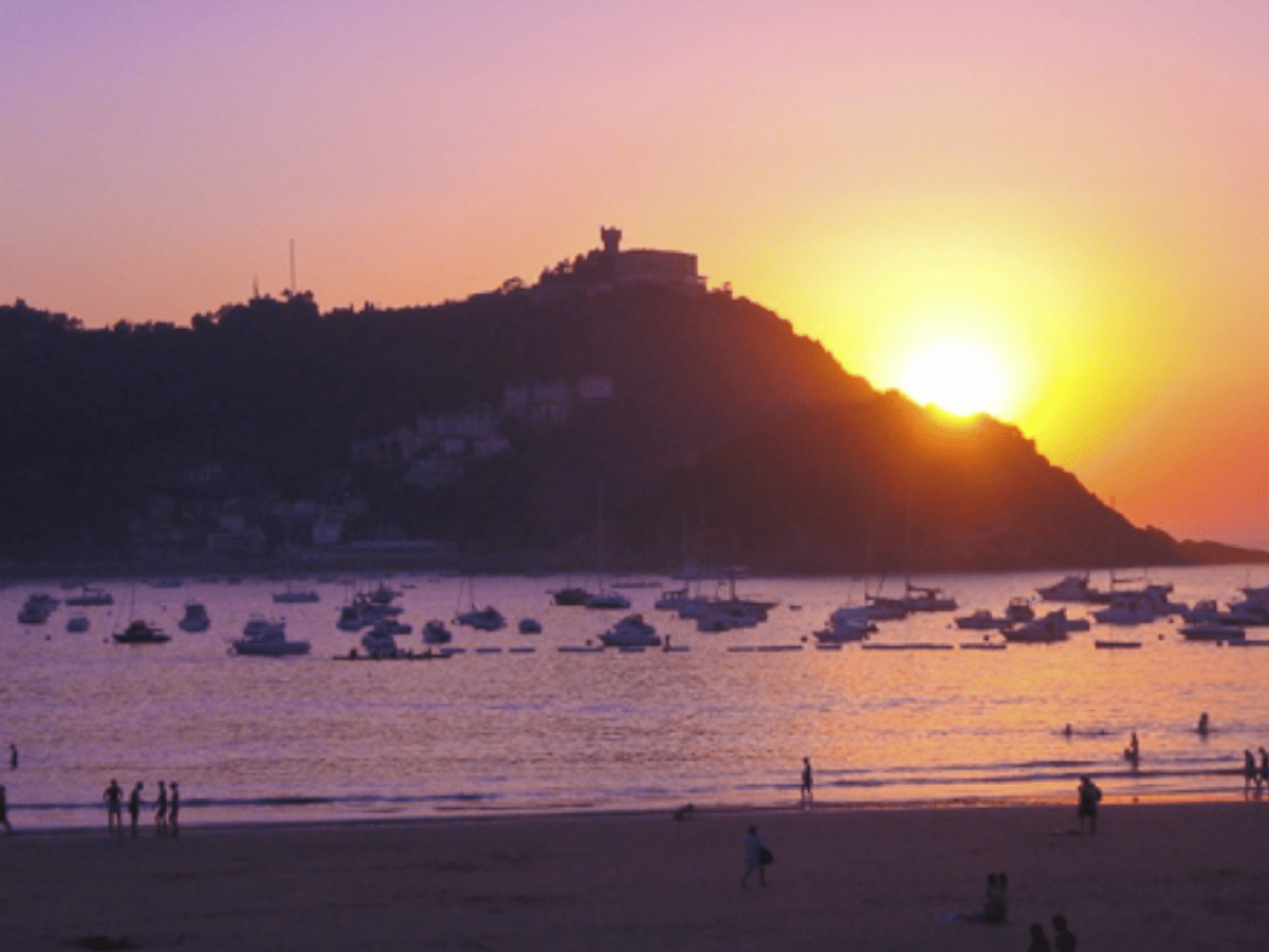 Sunset over a bay with boats and silhouettes of people on the beach.