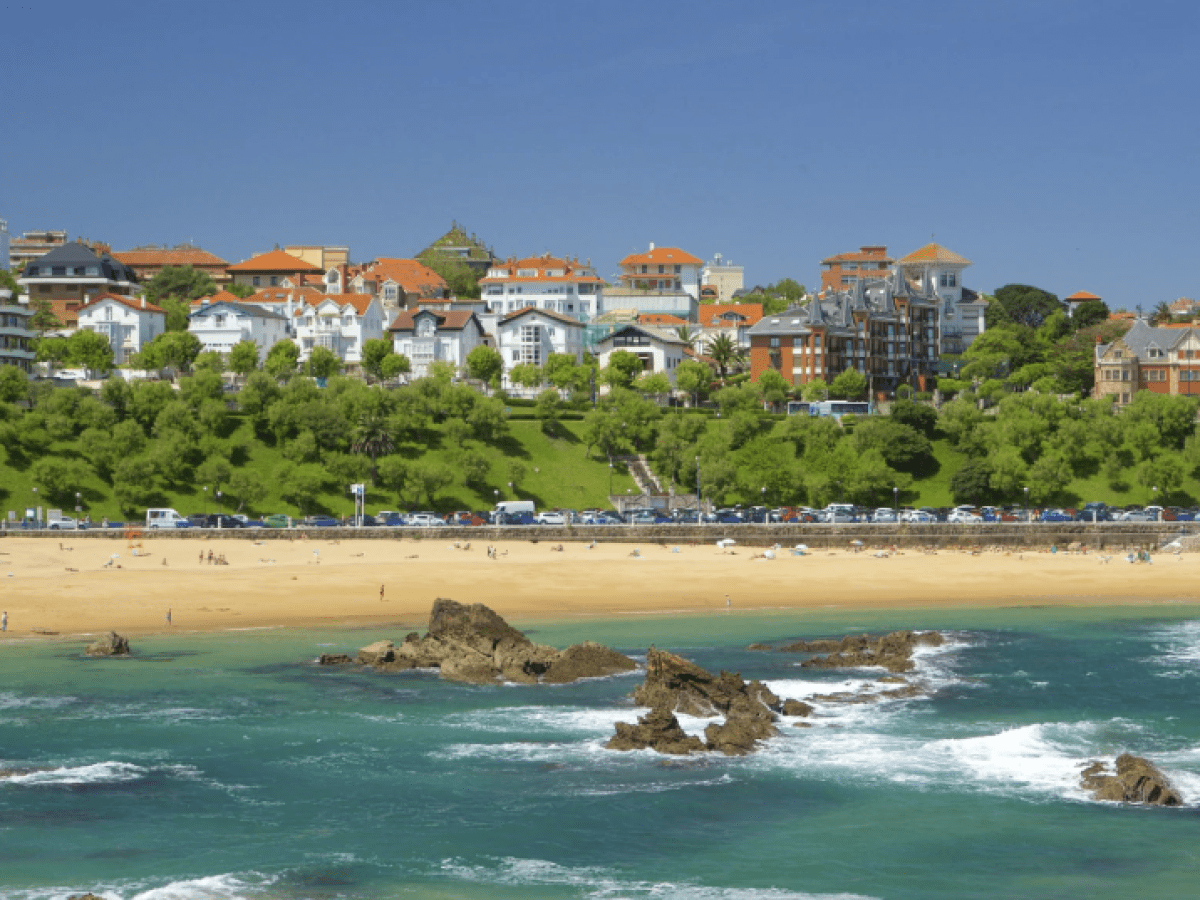 Beach with rock formations, green hills, and a town with colorful buildings under a clear blue sky.
