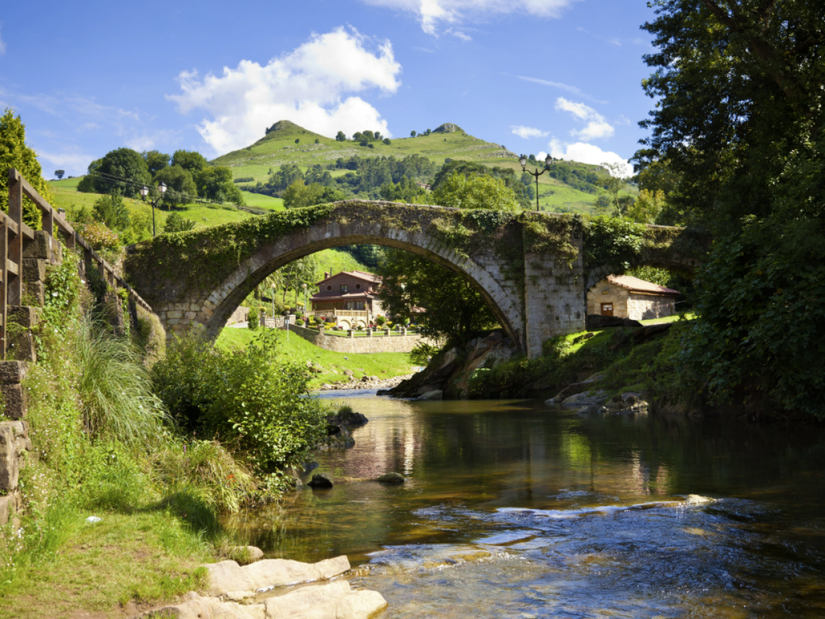 Stone bridge over a river with hills and a house in the background under a blue sky.