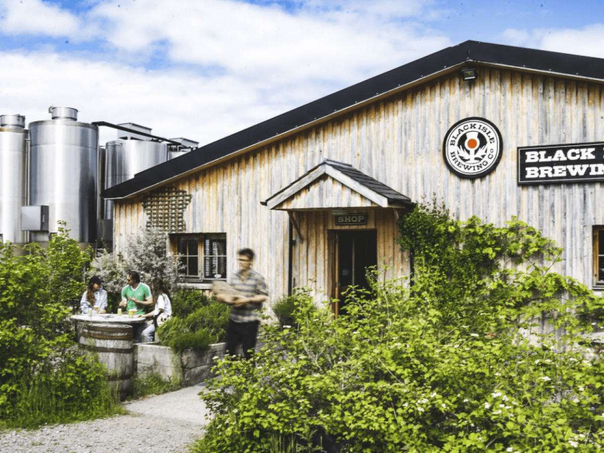 Brewery exterior with people sitting outside, surrounded by greenery and metal tanks.