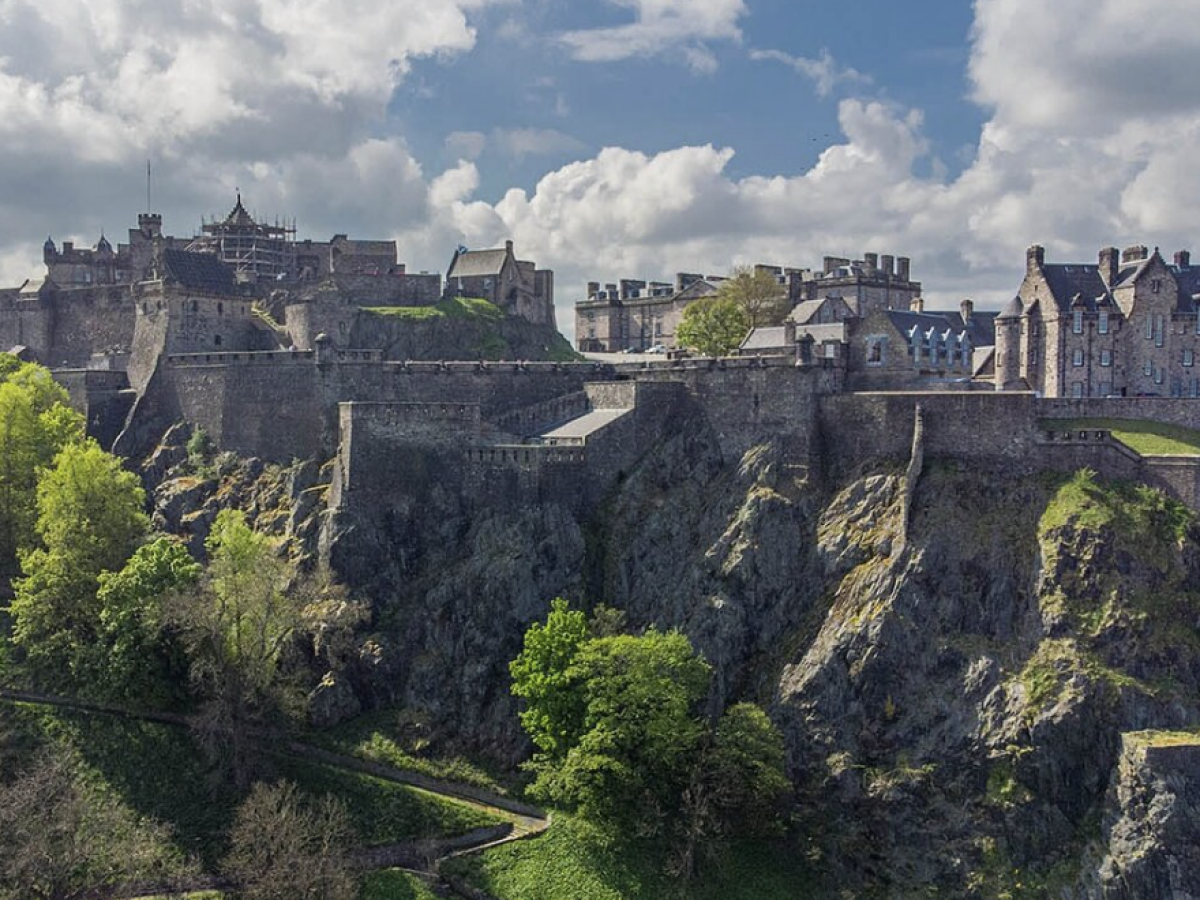 Medieval castle with walls on a rocky hill surrounded by green trees under a cloudy sky.