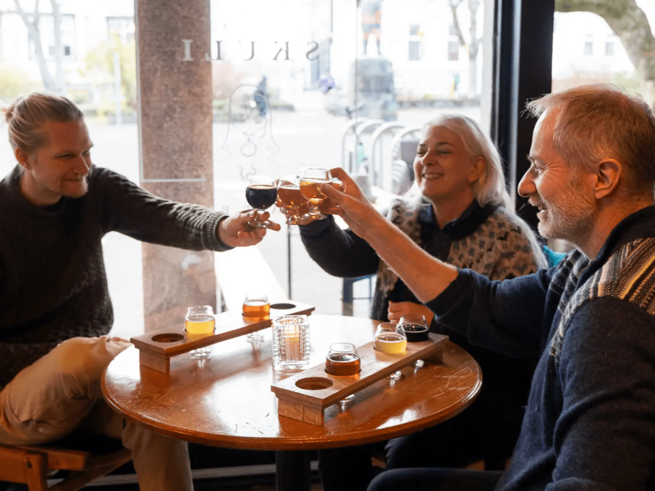 Three people at a cafe table clinking glasses with beer samples on trays.