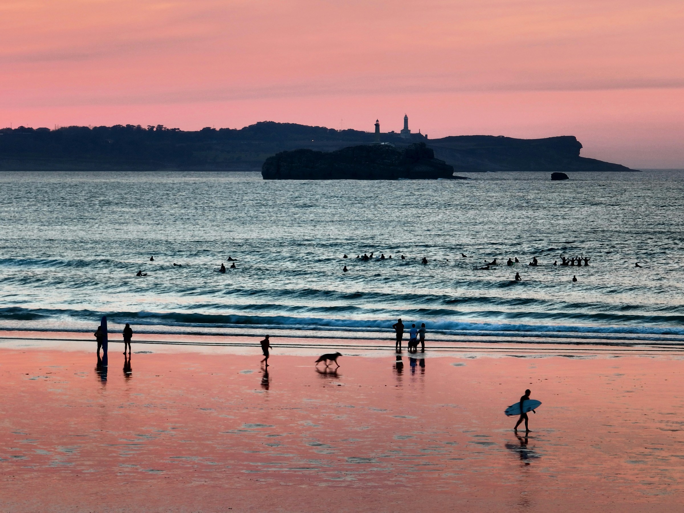 People and a surfer on a beach at sunset with calm waves and a distant lighthouse silhouette.