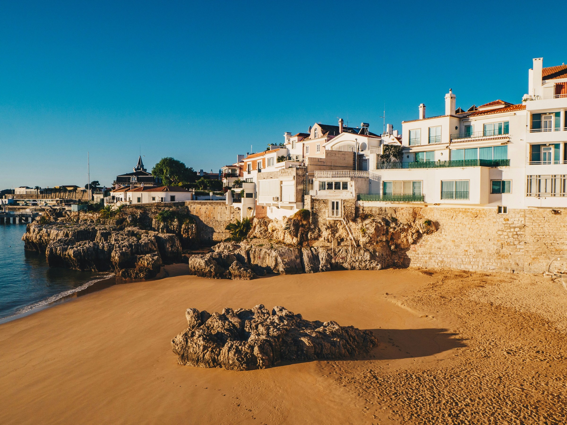 Coastal town with sandy beach, rocky shore, and buildings under a clear blue sky.