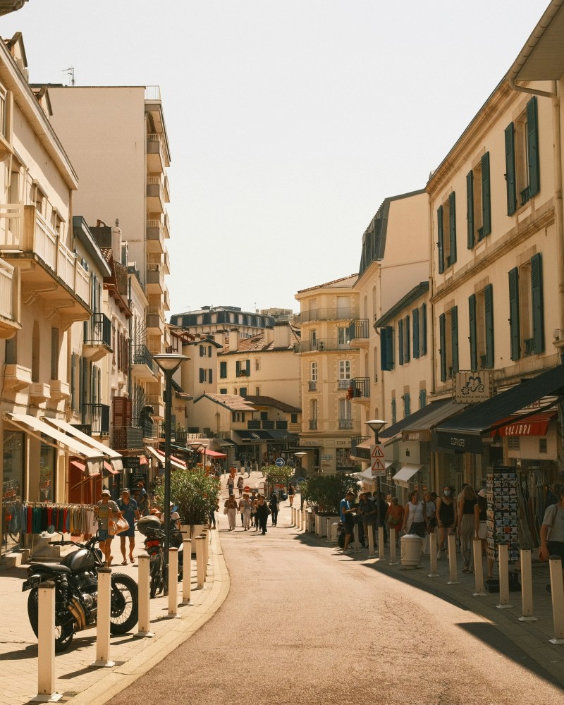 Sunny street scene with people, shops, and parked motorcycles in a European town.