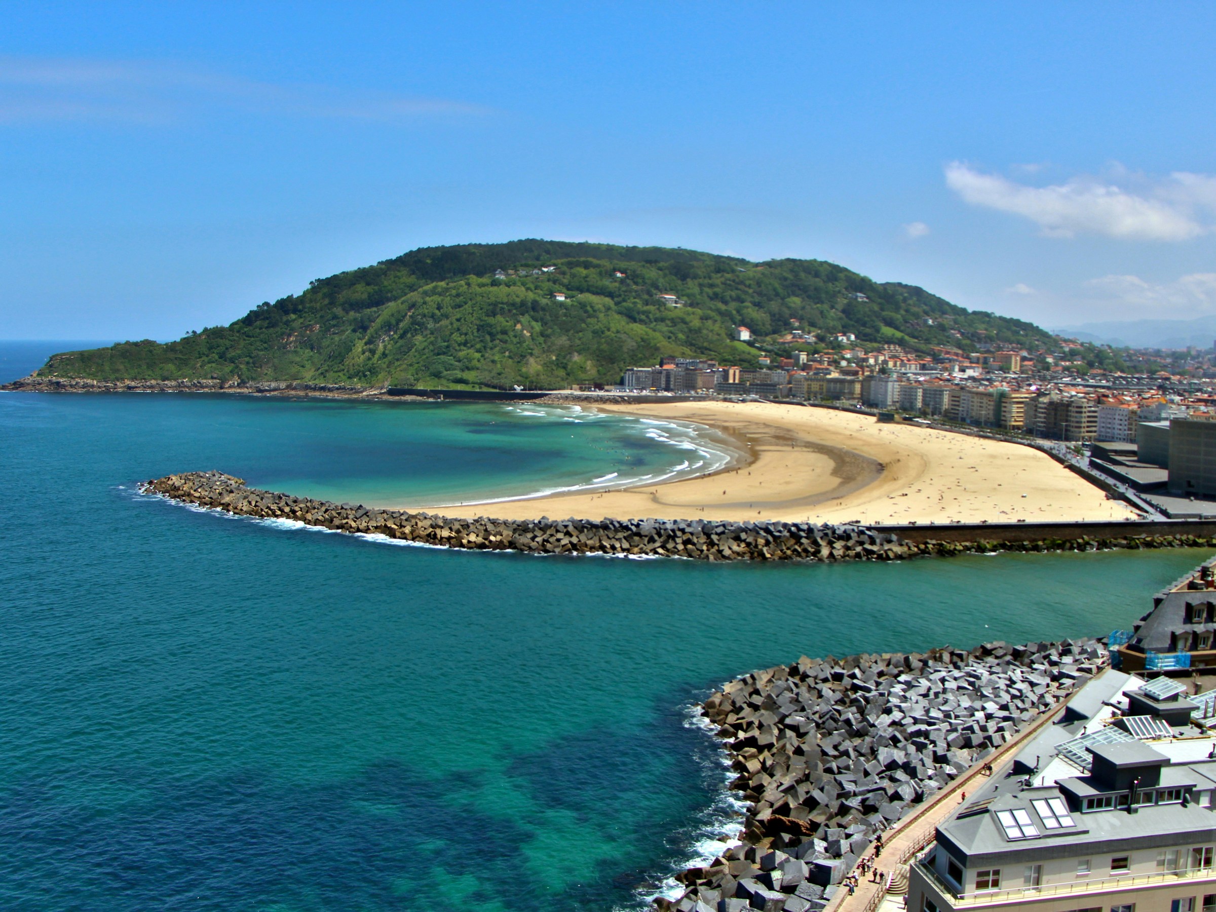 Coastal cityscape with sandy beach, green hillside, and turquoise sea under a blue sky.