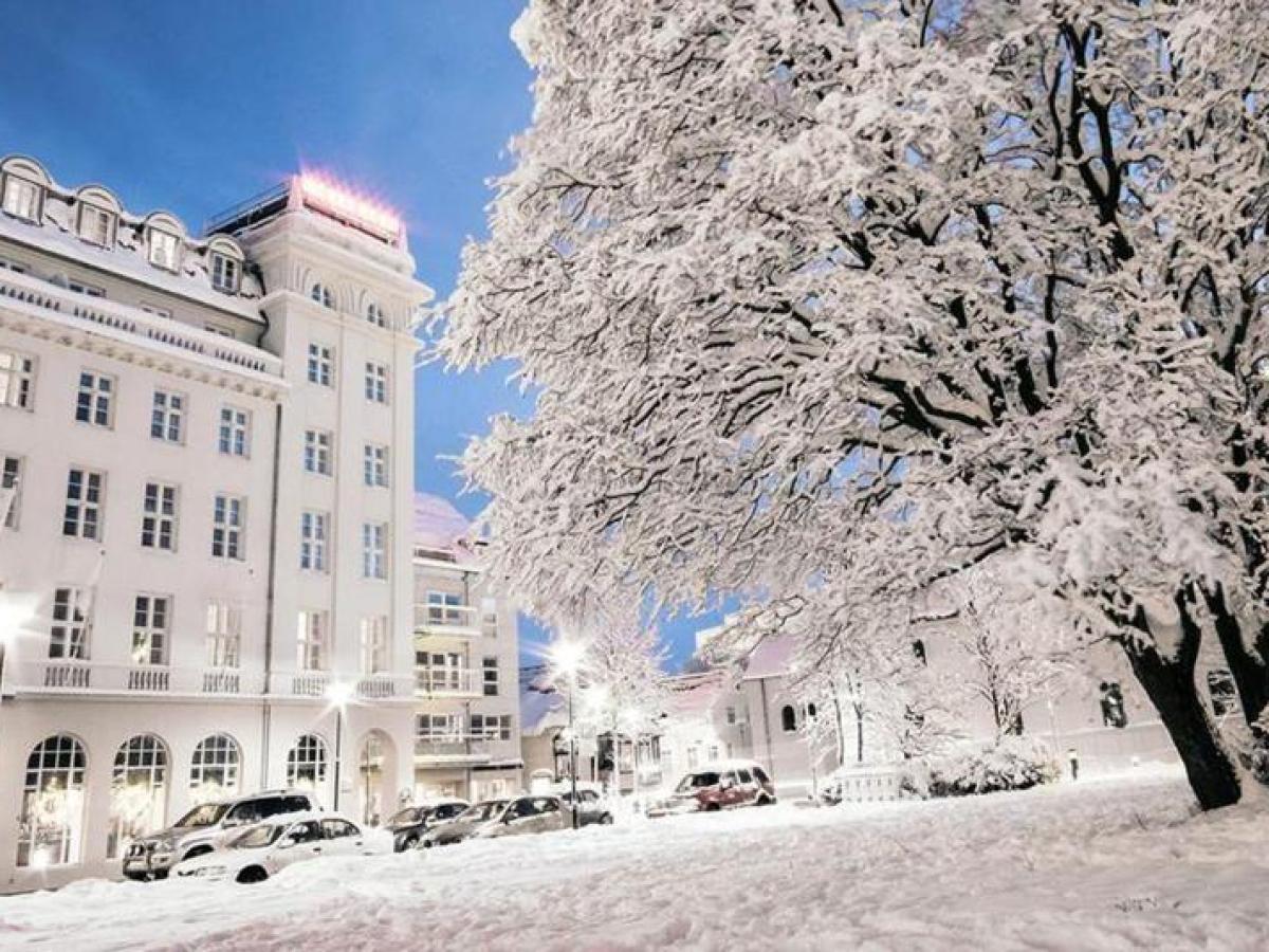 Snow-covered street with a building and trees under a clear blue sky.
