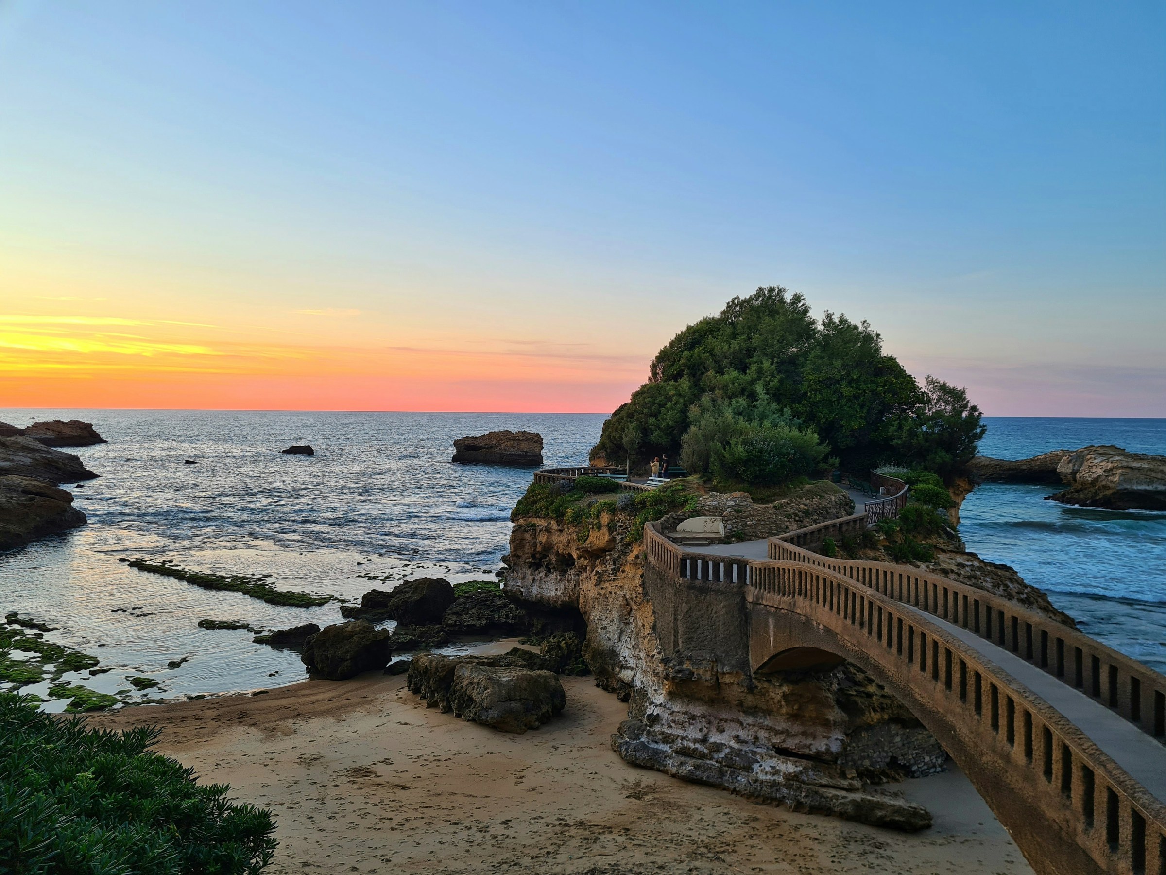 Sunset over ocean with a small green island connected by a bridge, rocky coastline in the foreground.