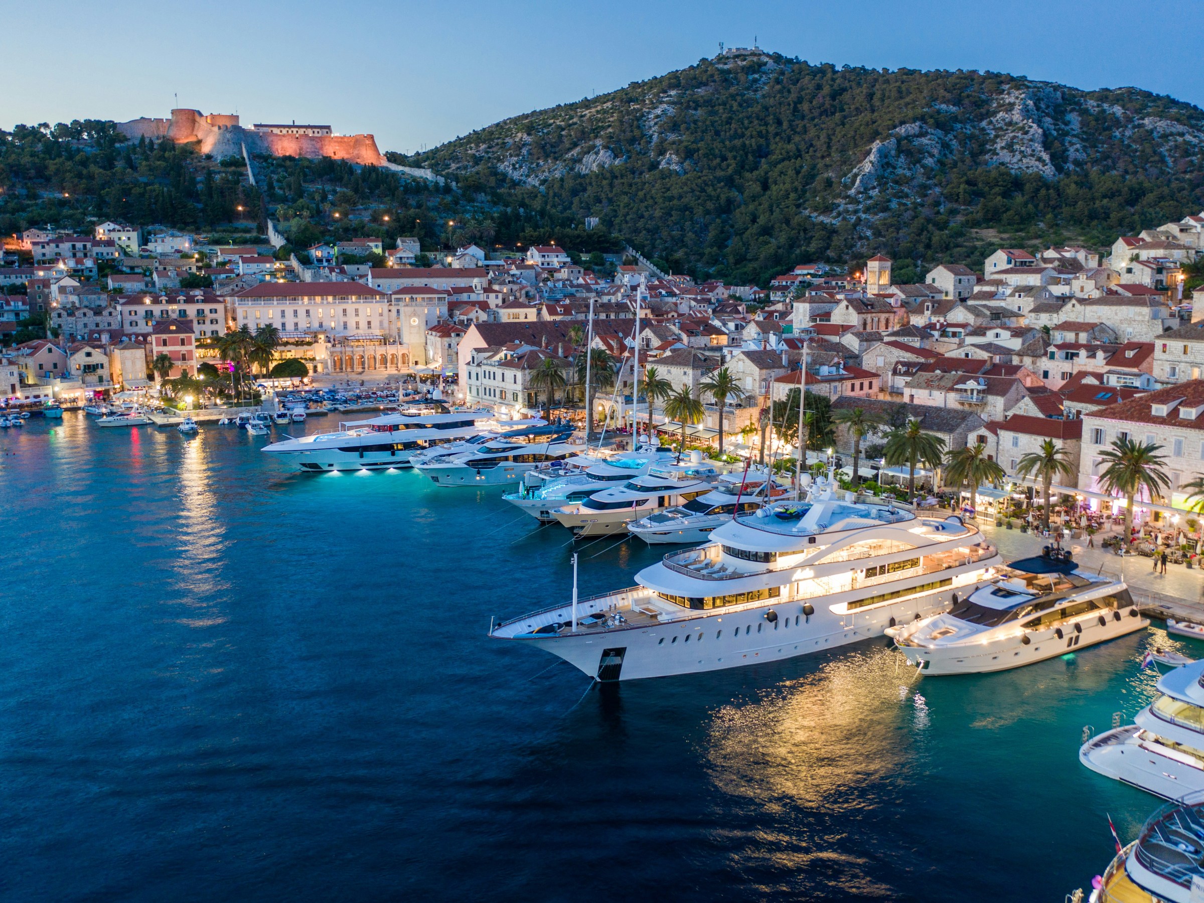Scenic harbor at dusk with yachts and hillside buildings under a lit-up fortress.