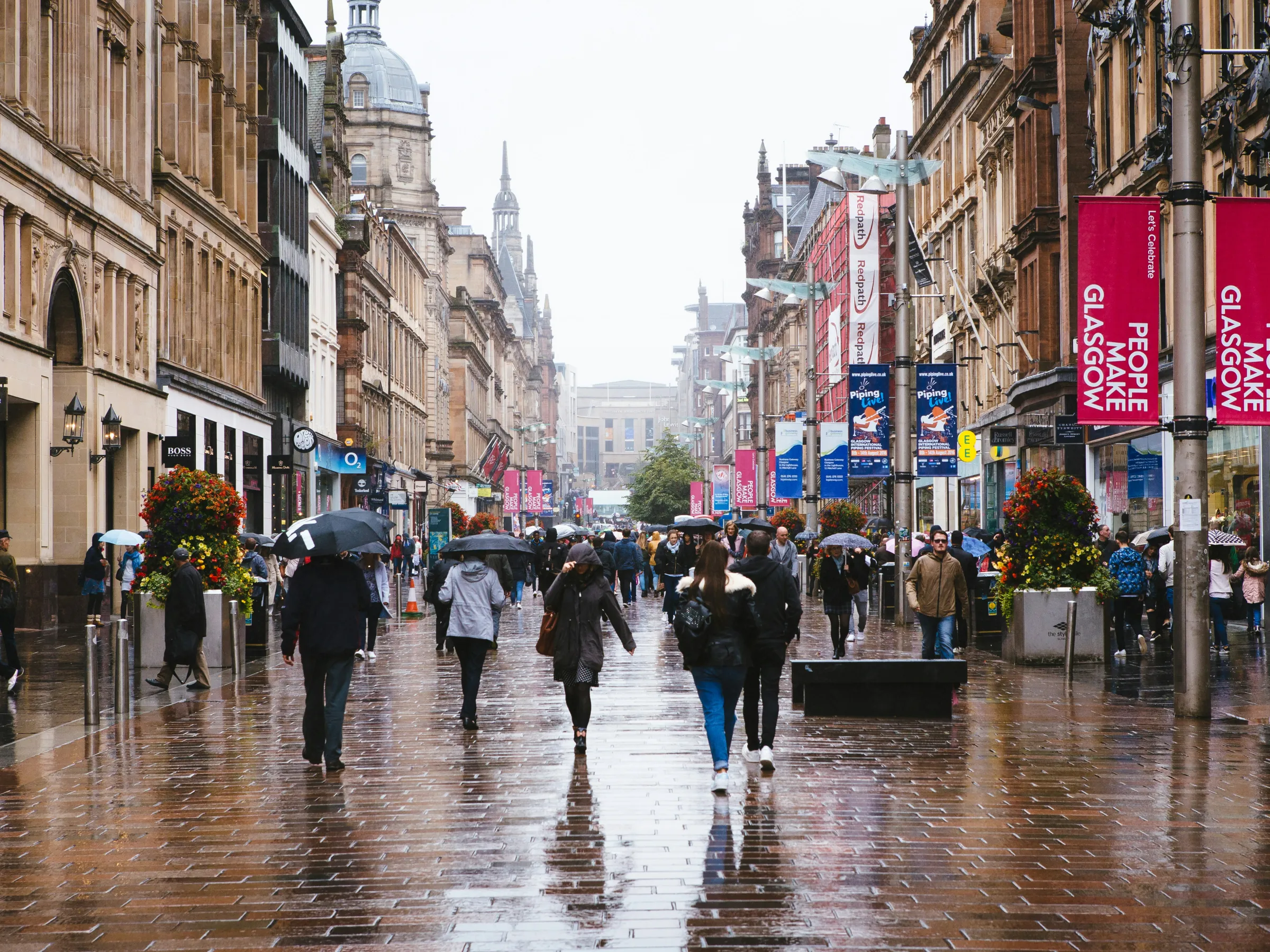 A wet city street with people carrying umbrellas, surrounded by historic buildings and colorful banners.