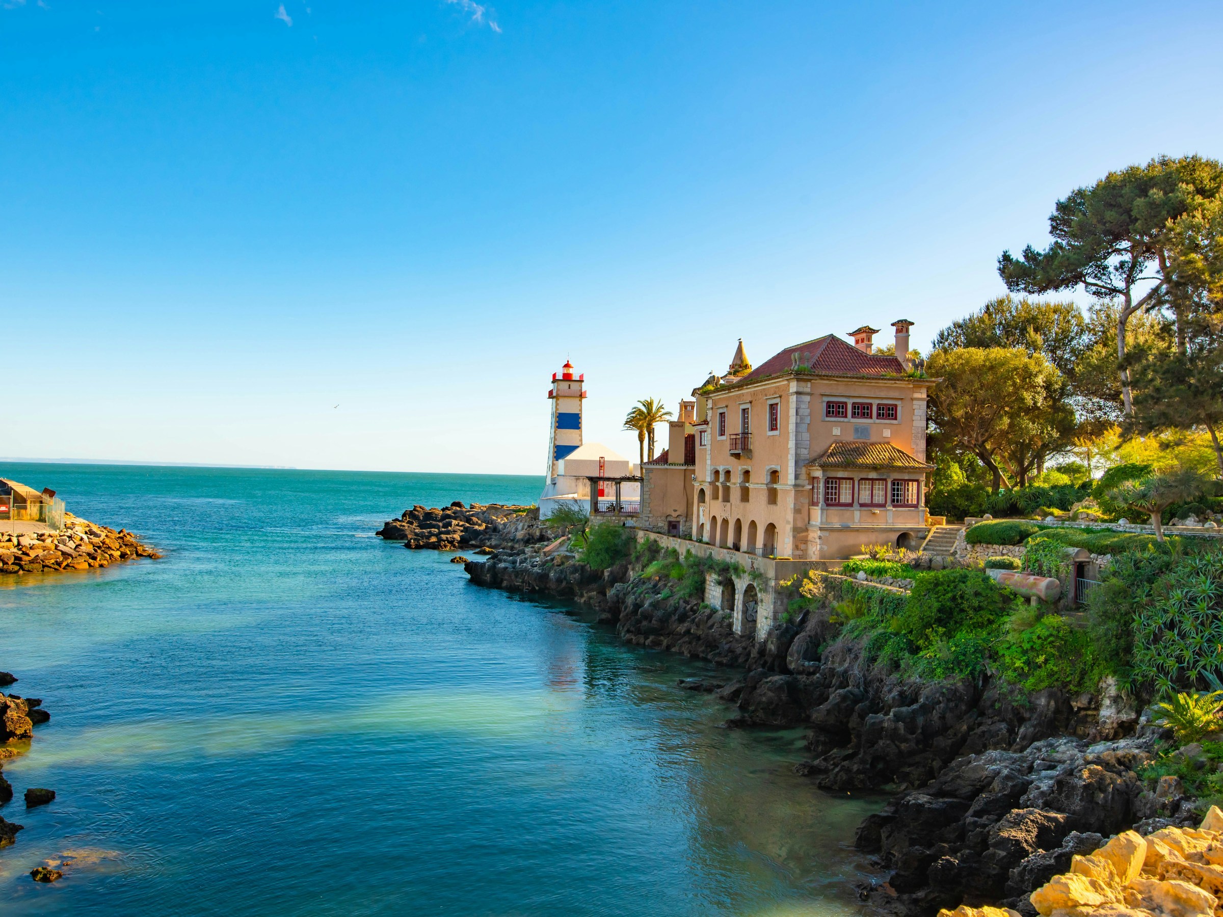 Coastal mansion with adjacent lighthouse, surrounded by greenery and blue ocean under clear sky.