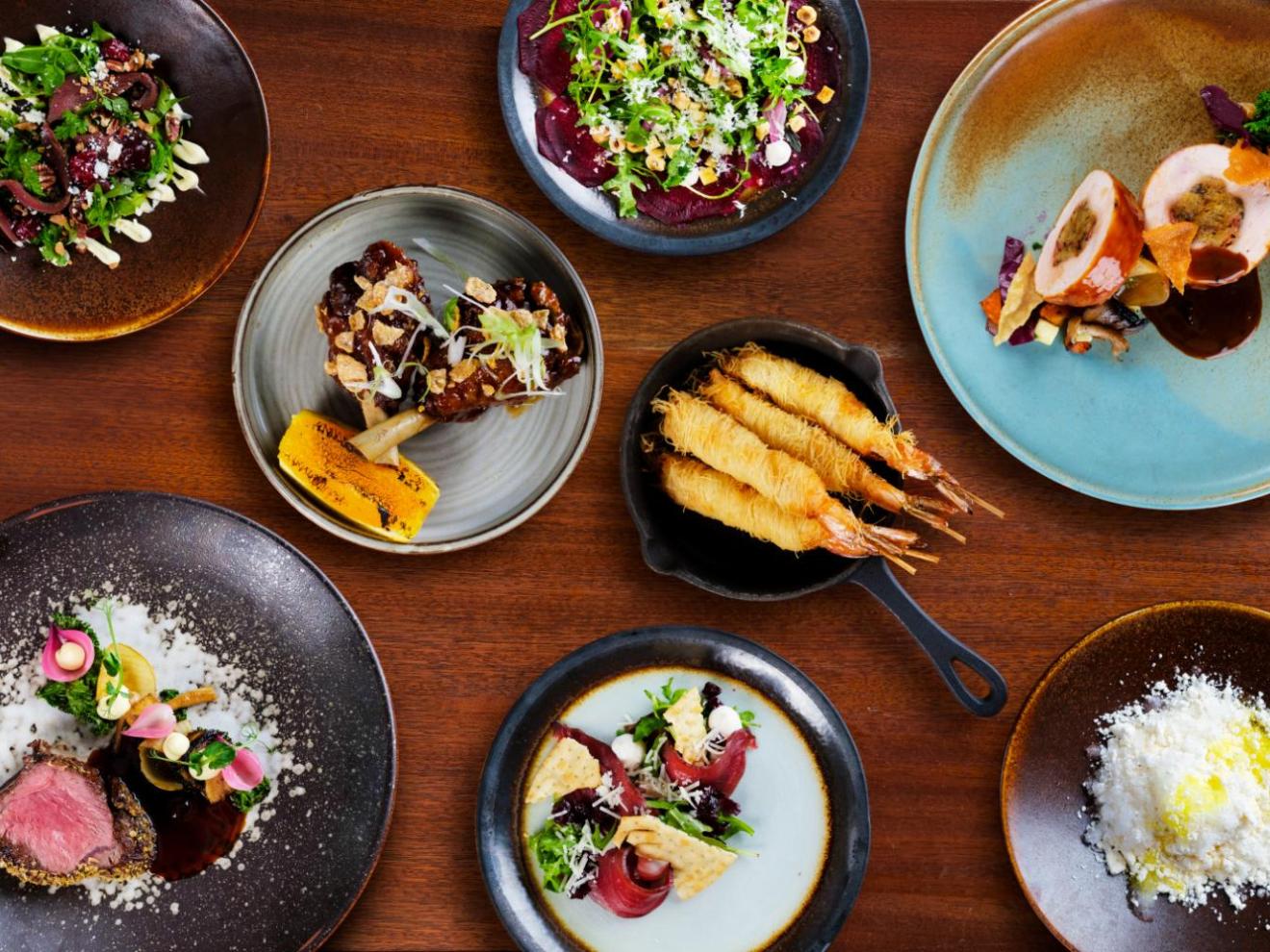 A variety of gourmet dishes plated on different colored plates on a wooden table.