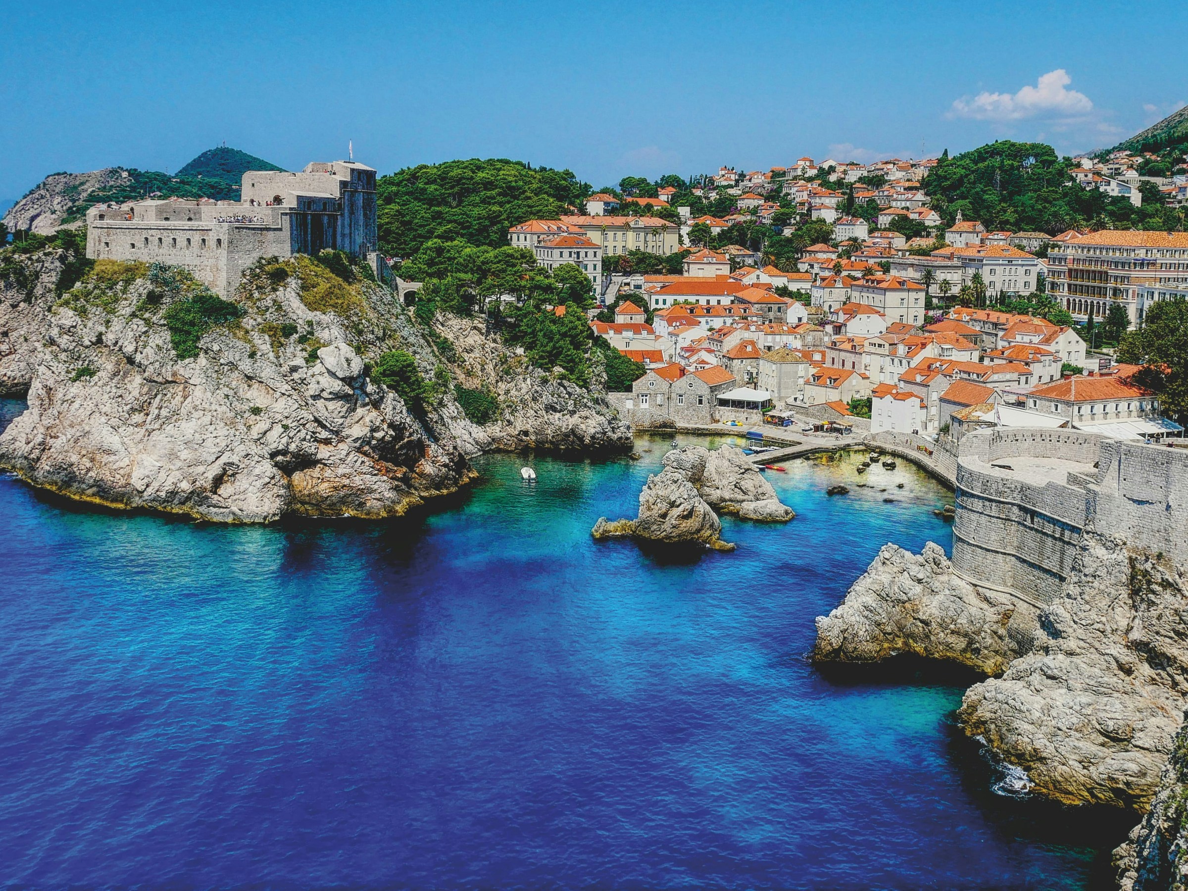 Scenic coastal city with red-roofed buildings and fortresses on a rocky shoreline against clear blue water.