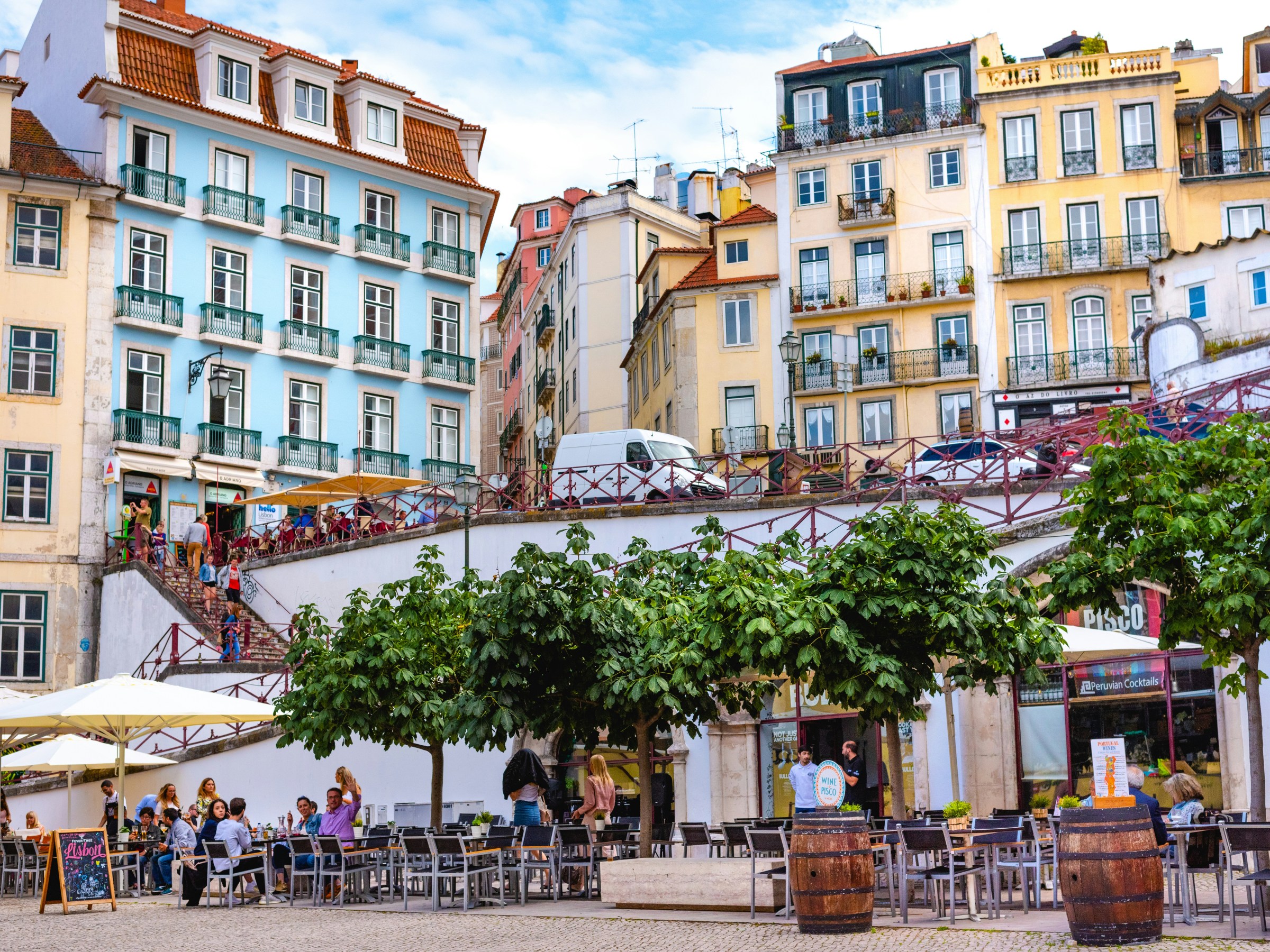 Outdoor café with people under trees, surrounded by colorful buildings in a hilly area.