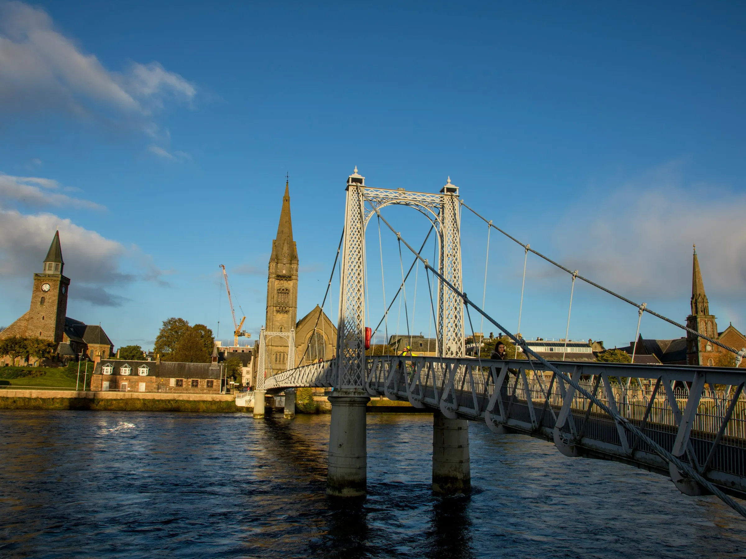 Suspension bridge over a river with church towers in the background under a clear blue sky.