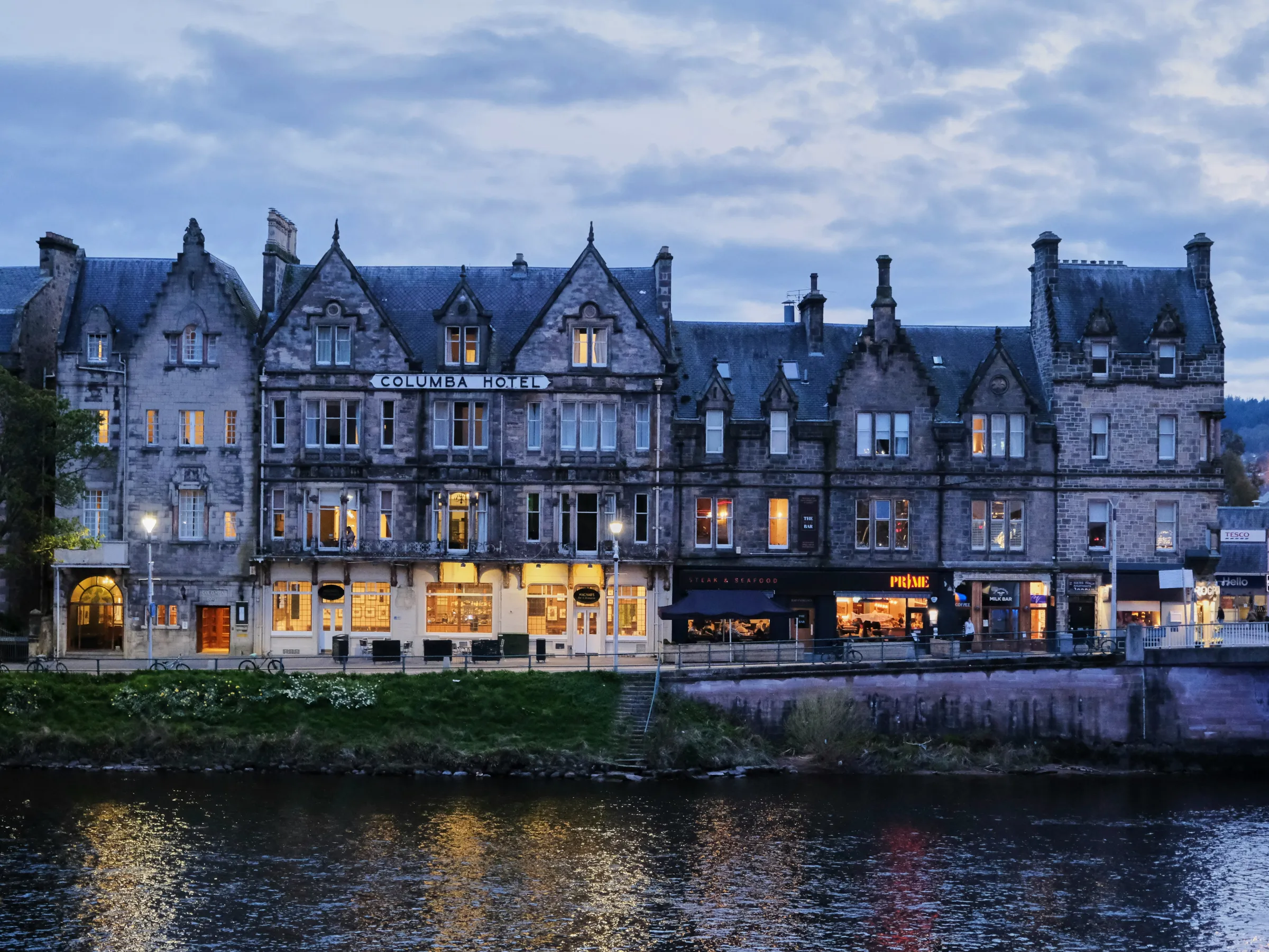 Historic stone building with lit windows by a river at dusk, labeled 'Columba Hotel'.