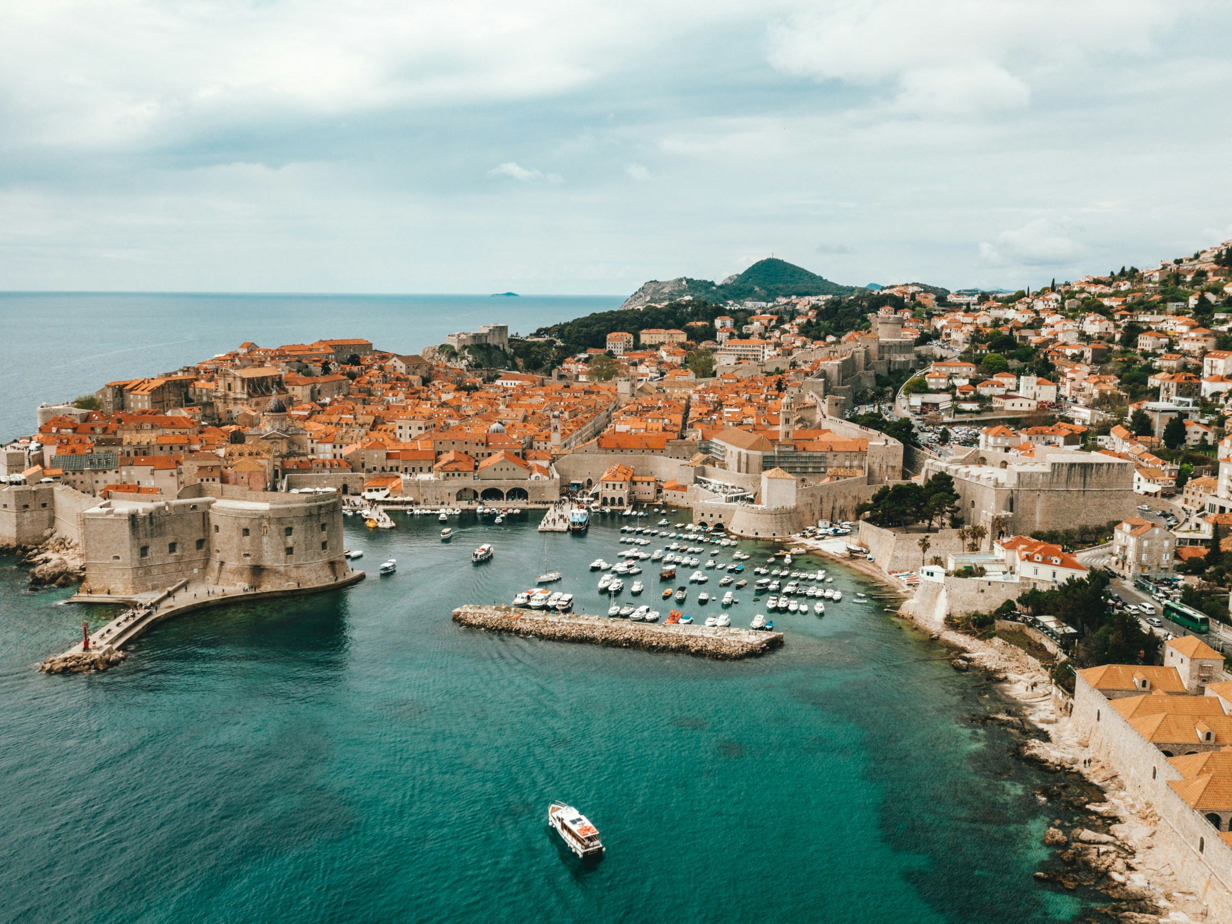 Aerial view of a historic coastal city with orange rooftops and a marina in clear blue water.
