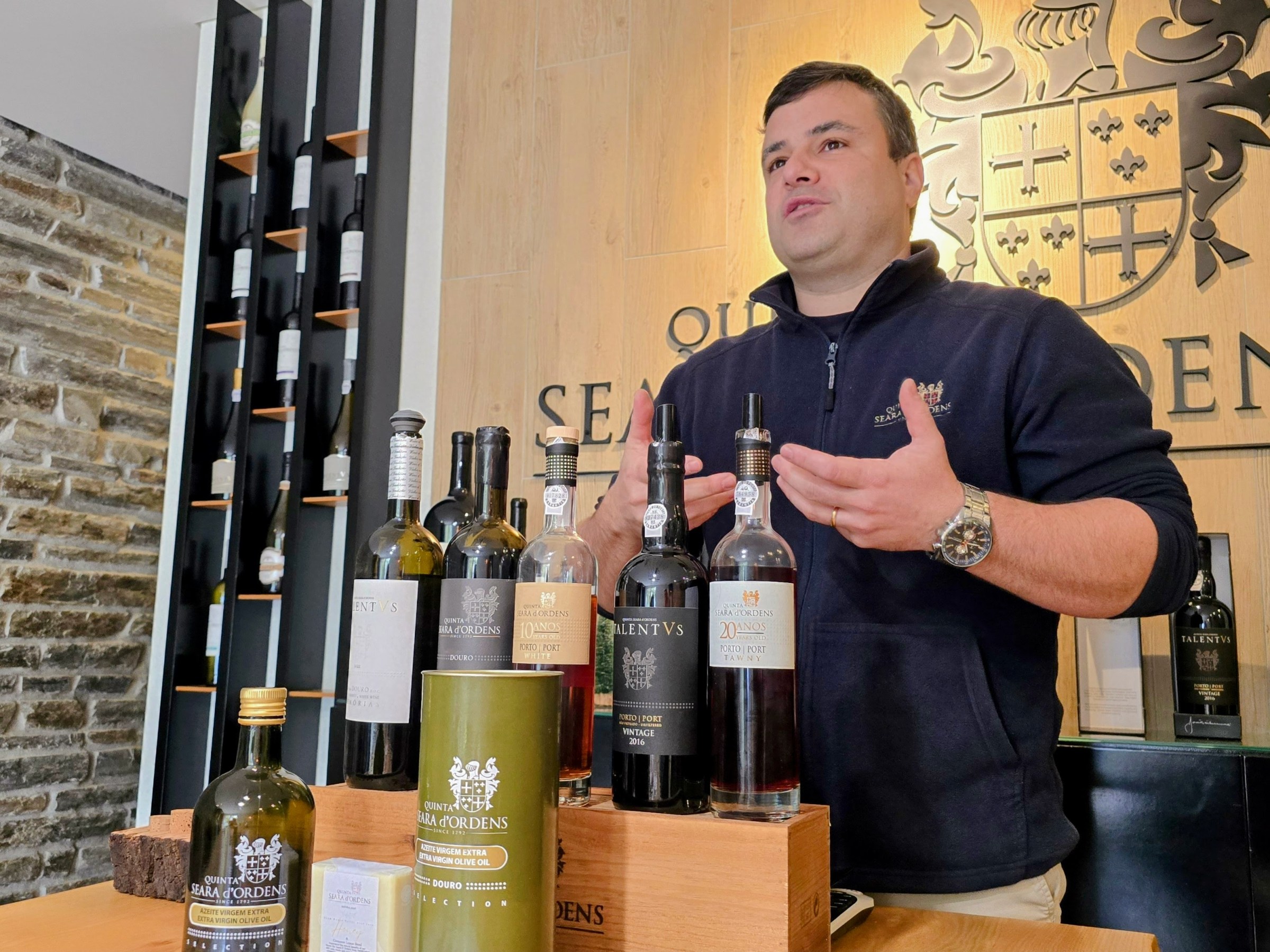 Man presenting wine bottles in a winery, with a decorative crest on the wall behind.