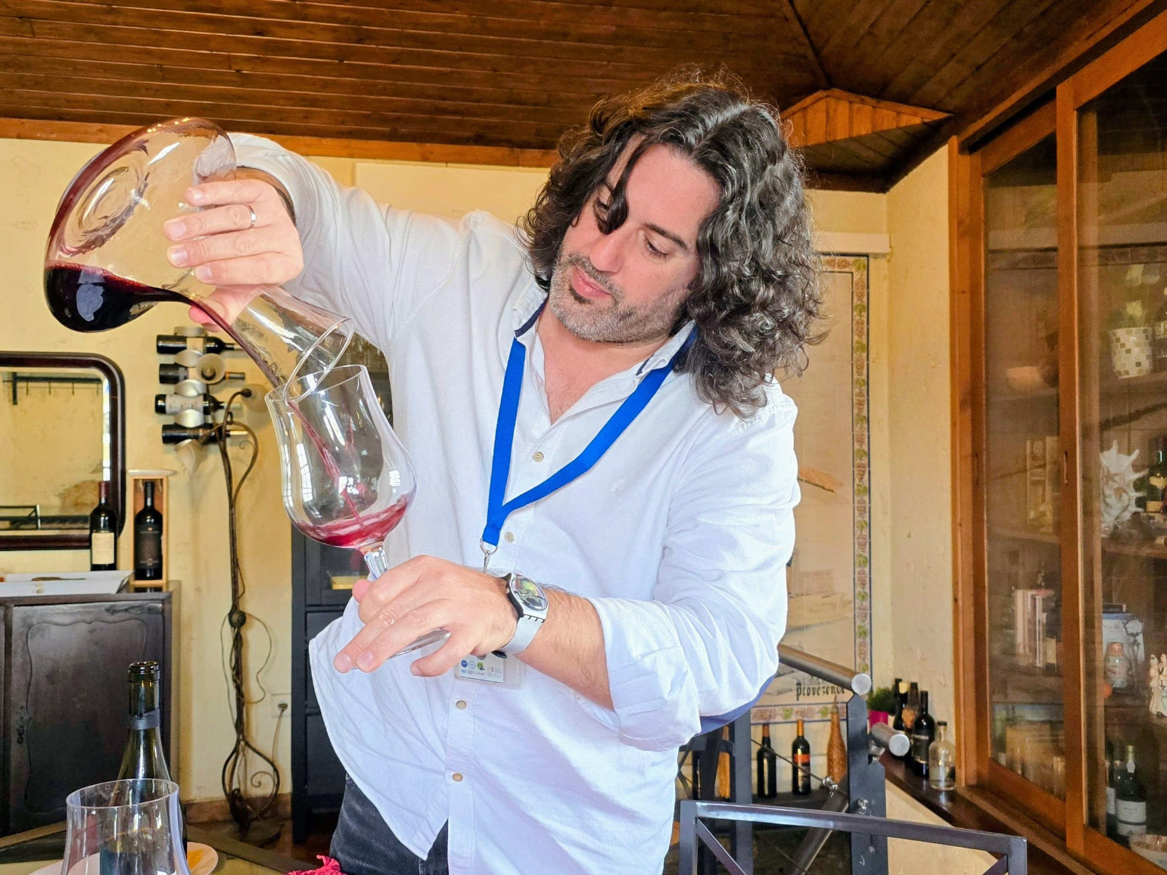 Person pouring red wine from decanter into glass in a cozy room.