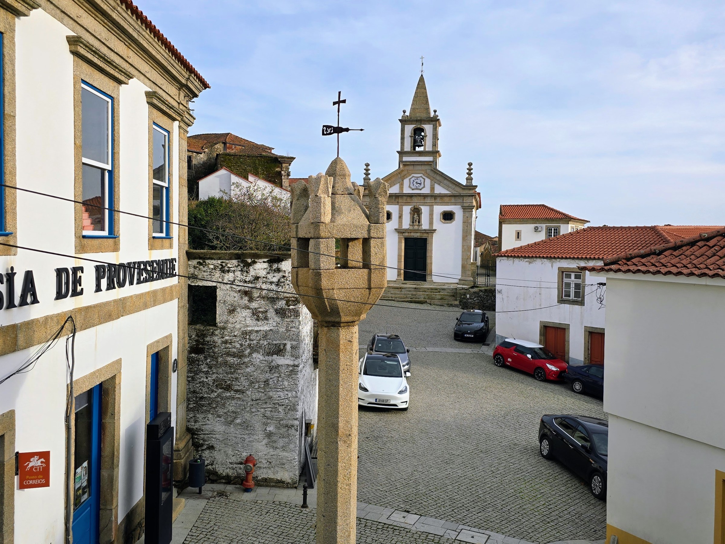 Stone cross in a cobblestone square with church and buildings on a sunny day.