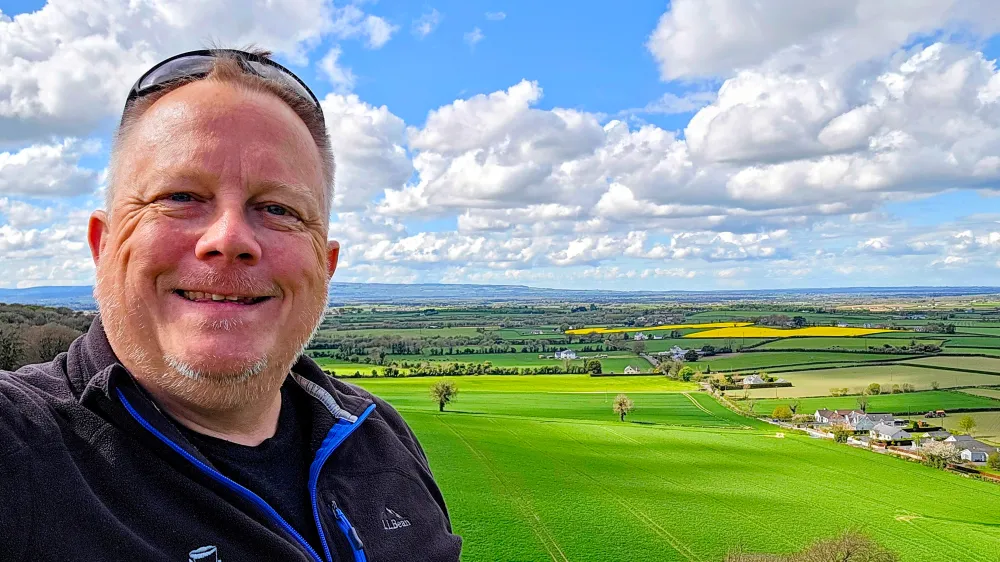 Person taking a selfie with green fields and cloudy sky in the background.