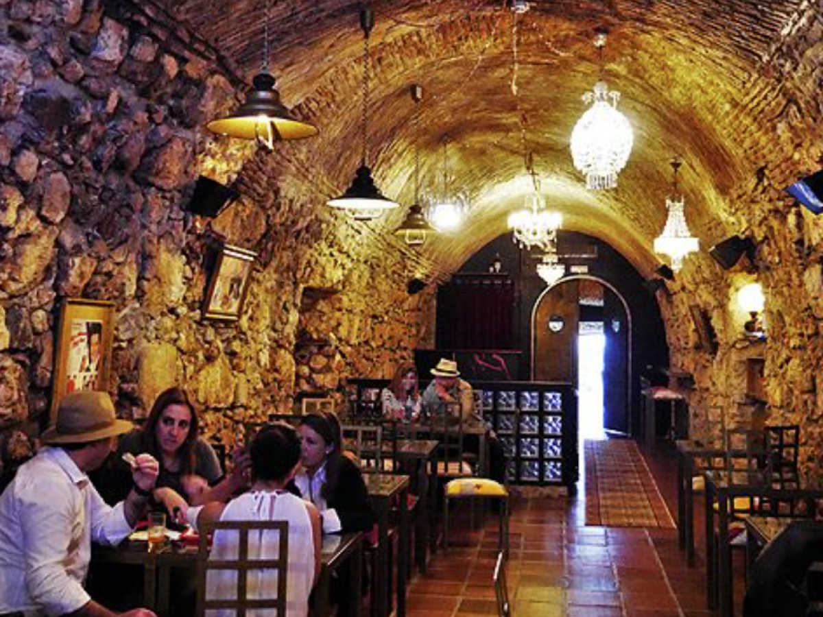 Rustic stone-walled café with chandeliers, diners seated at tables, arched ceiling, and warm lighting.